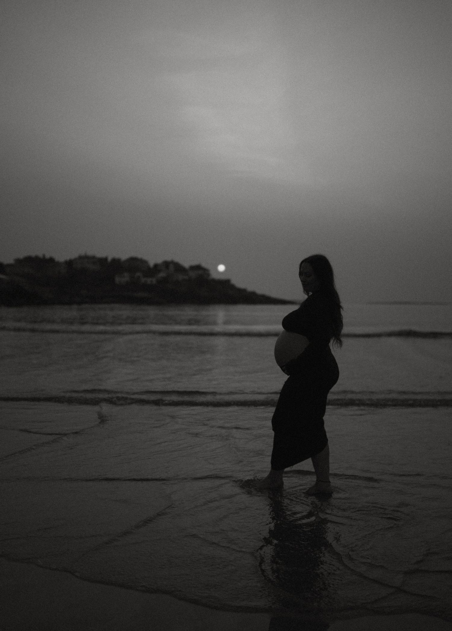 Black and white silhouette of pregnant woman standing in ocean waves at night with sun rising.