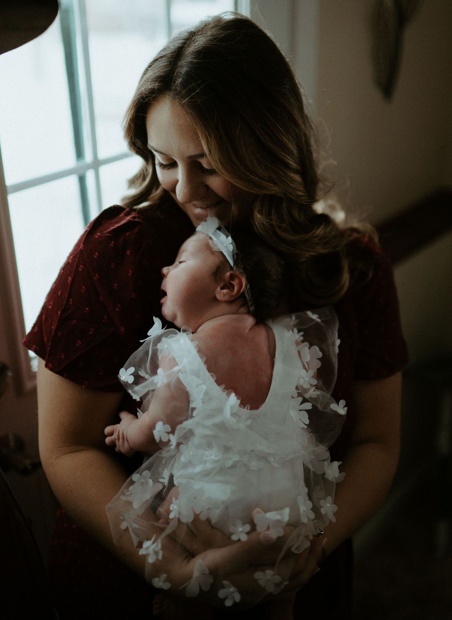 Loving mother in burgundy dress cradling newborn baby girl in white floral lace gown indoors.
