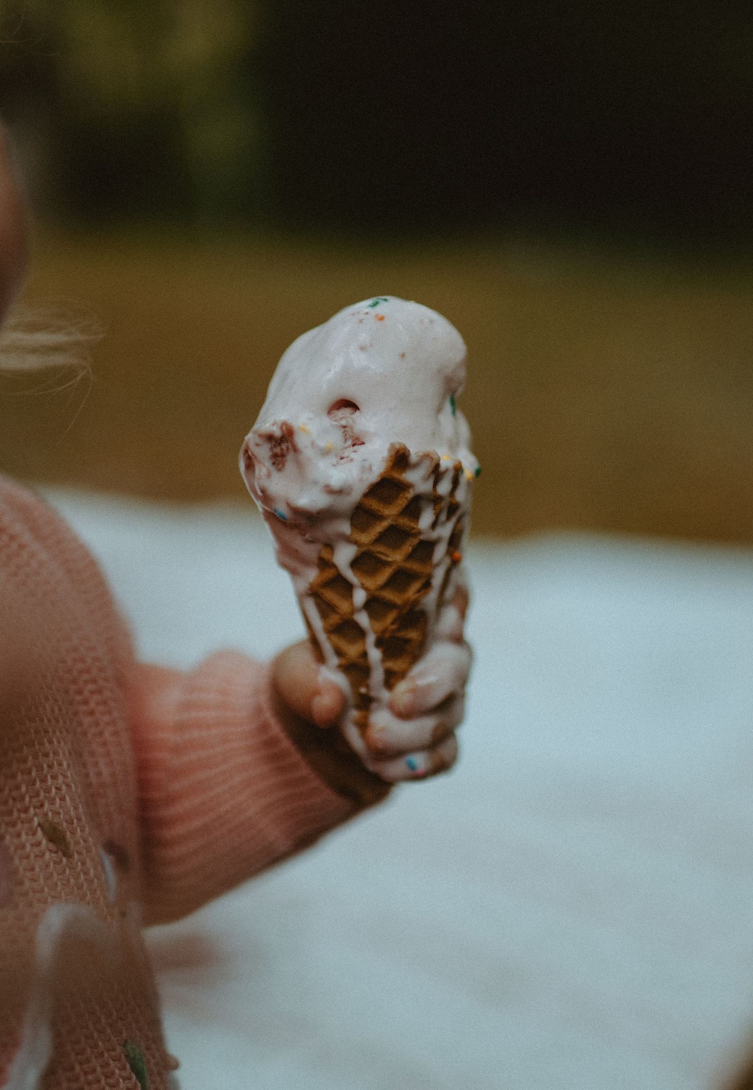 Close-up of a child holding a melting vanilla ice cream waffle cone with sprinkles outdoors.