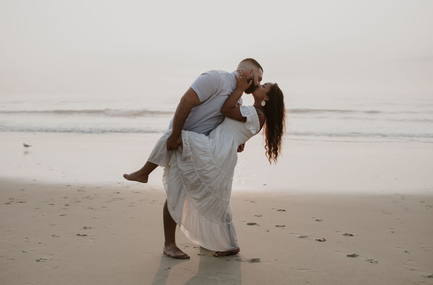Man dipping woman in white dress for a romantic kiss on the beach with ocean waves behind them.