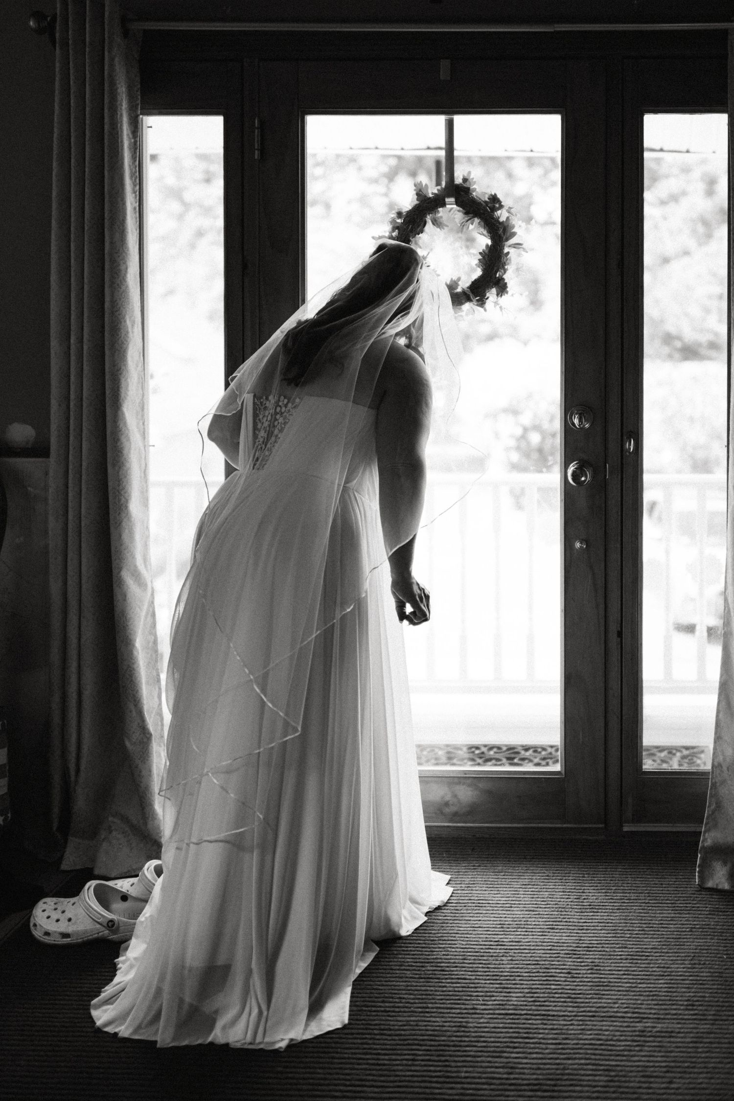 Silhouette of bride in flowing wedding gown and floral crown veil standing by glass doors in black and white.