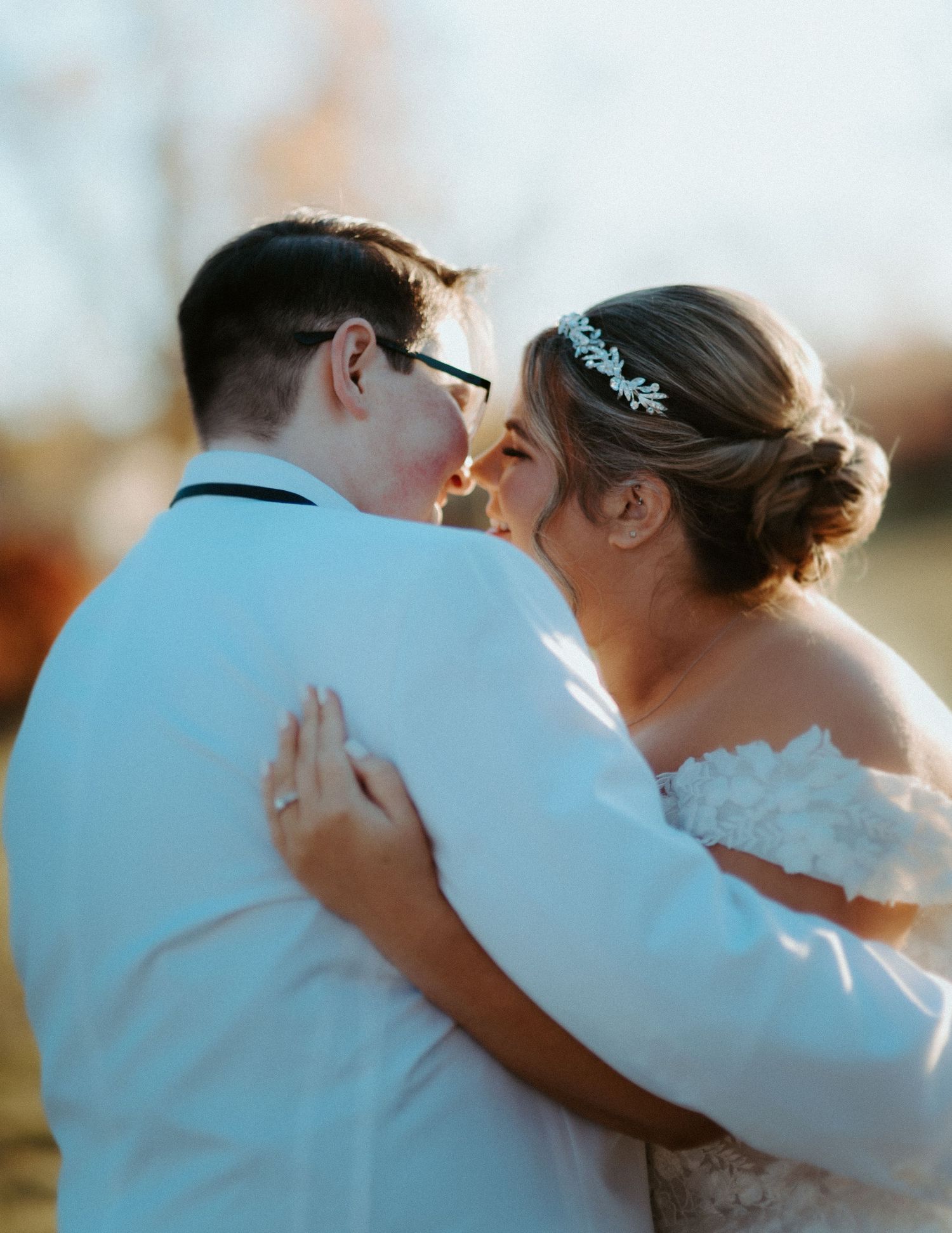 Brides nearly kissing outdoors in warm sunset light, one bride wearing lace gown and headpiece.