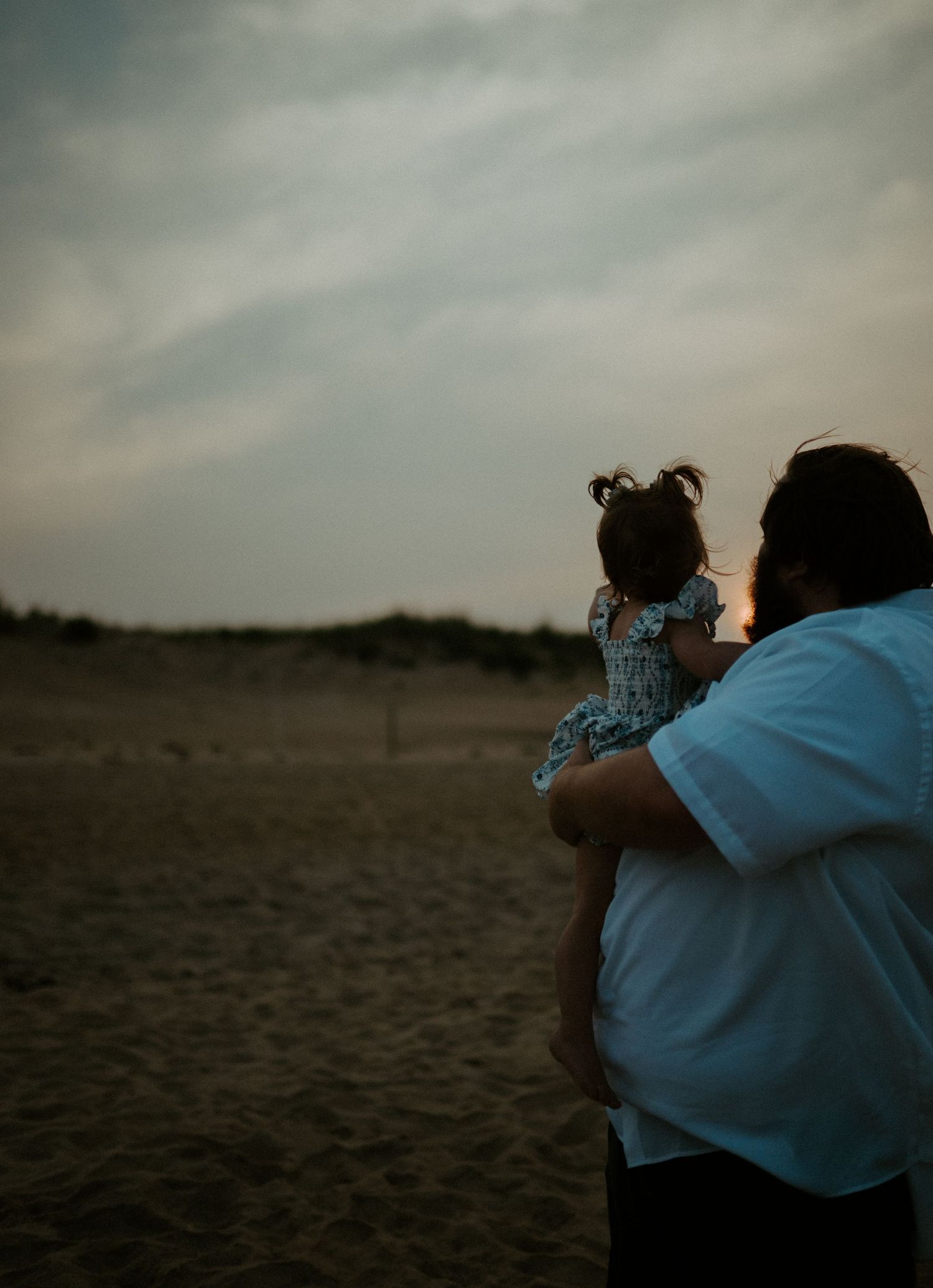 Father holds his young daughter while gazing at a dramatic cloudy sunset over sandy dunes.