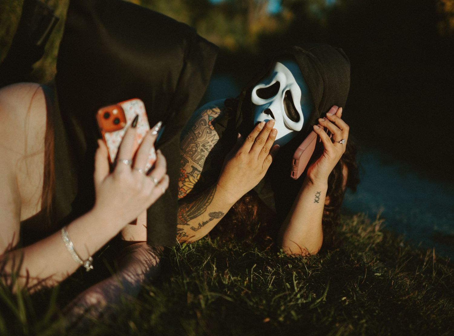 Two people in Ghostface Halloween costumes lying on grass talking on phones.