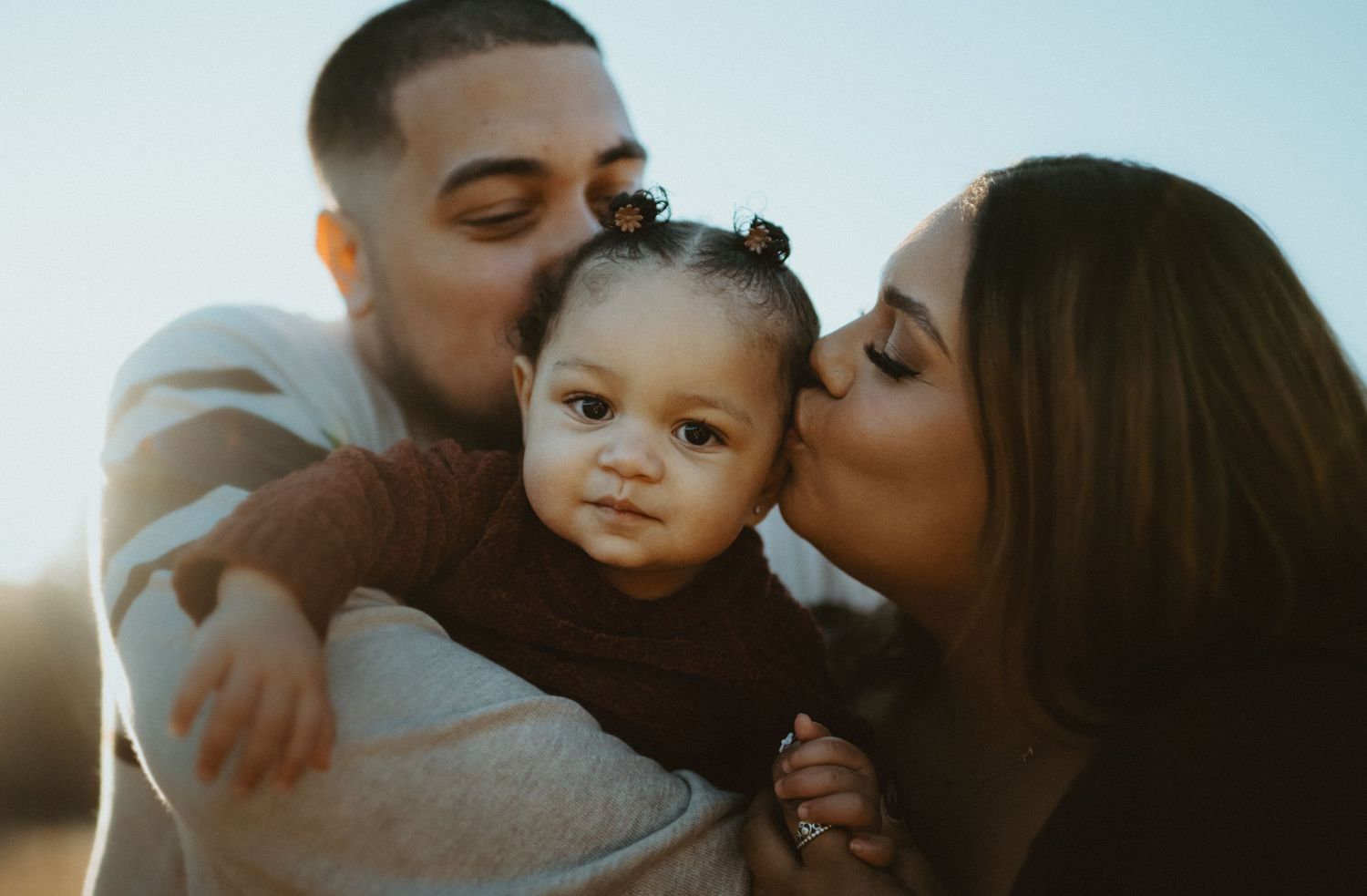 Parents kissing their baby girl on cheeks in golden sunset light outdoors.