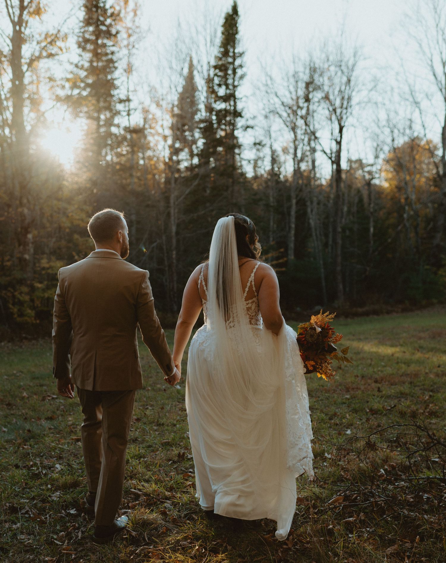 Bride and groom holding hands walking toward sunlit autumn forest with fall bouquet.