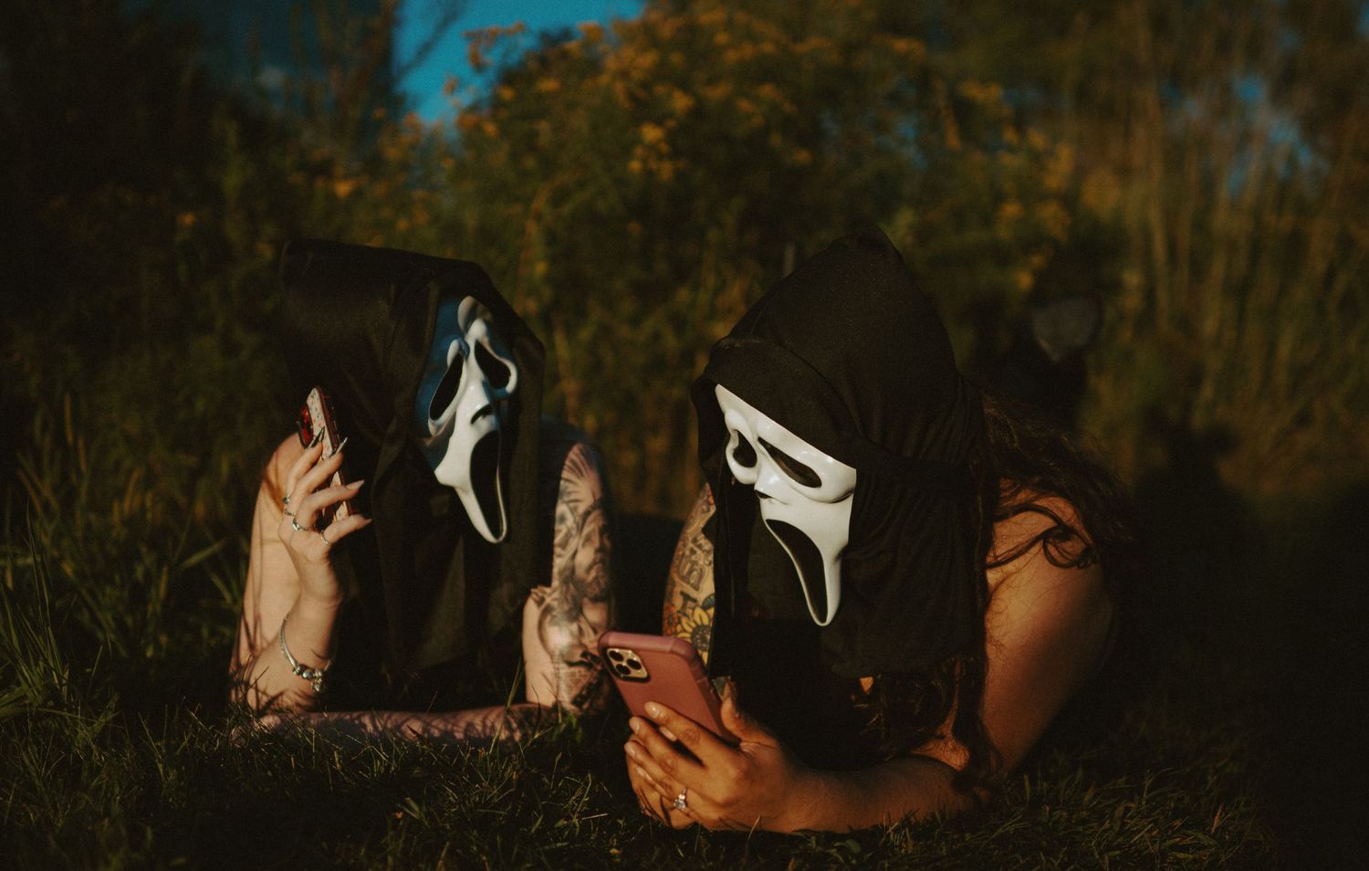 Two people wearing Ghostface Halloween masks lying in grass looking at phones