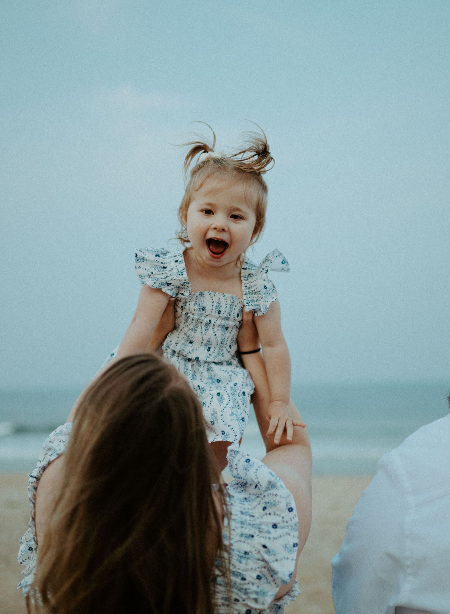 Joyful toddler girl in floral dress laughs with delight as mother lifts her on the beach.