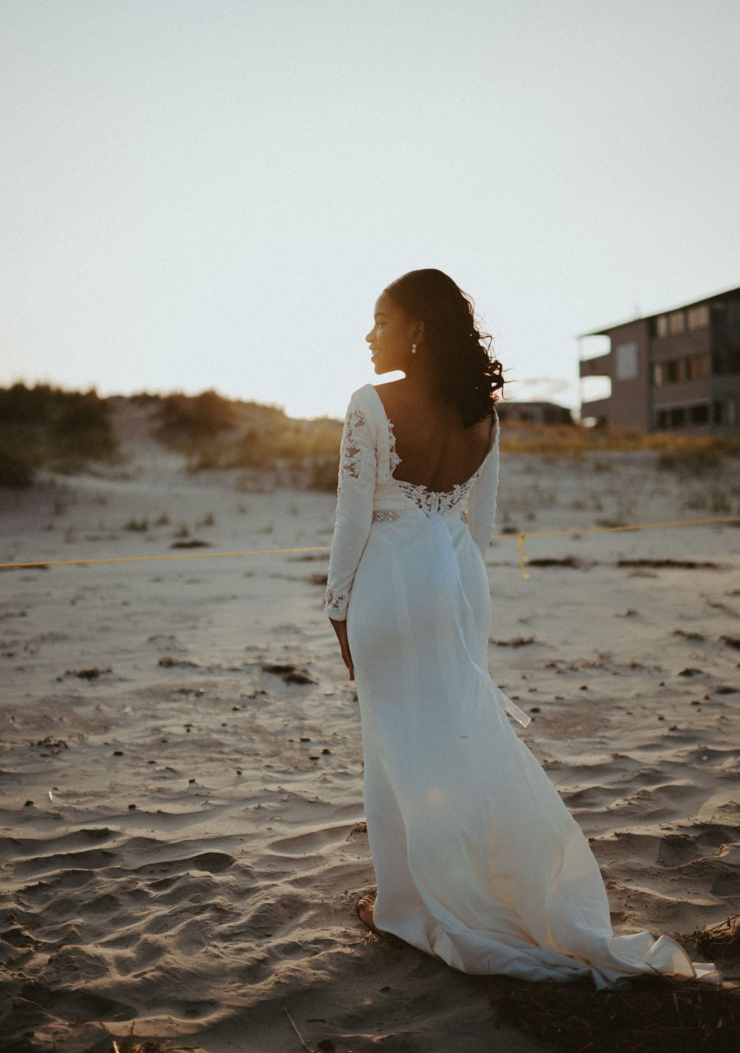 Bride in long-sleeve lace backless wedding gown walks on sandy beach at golden hour sunset.