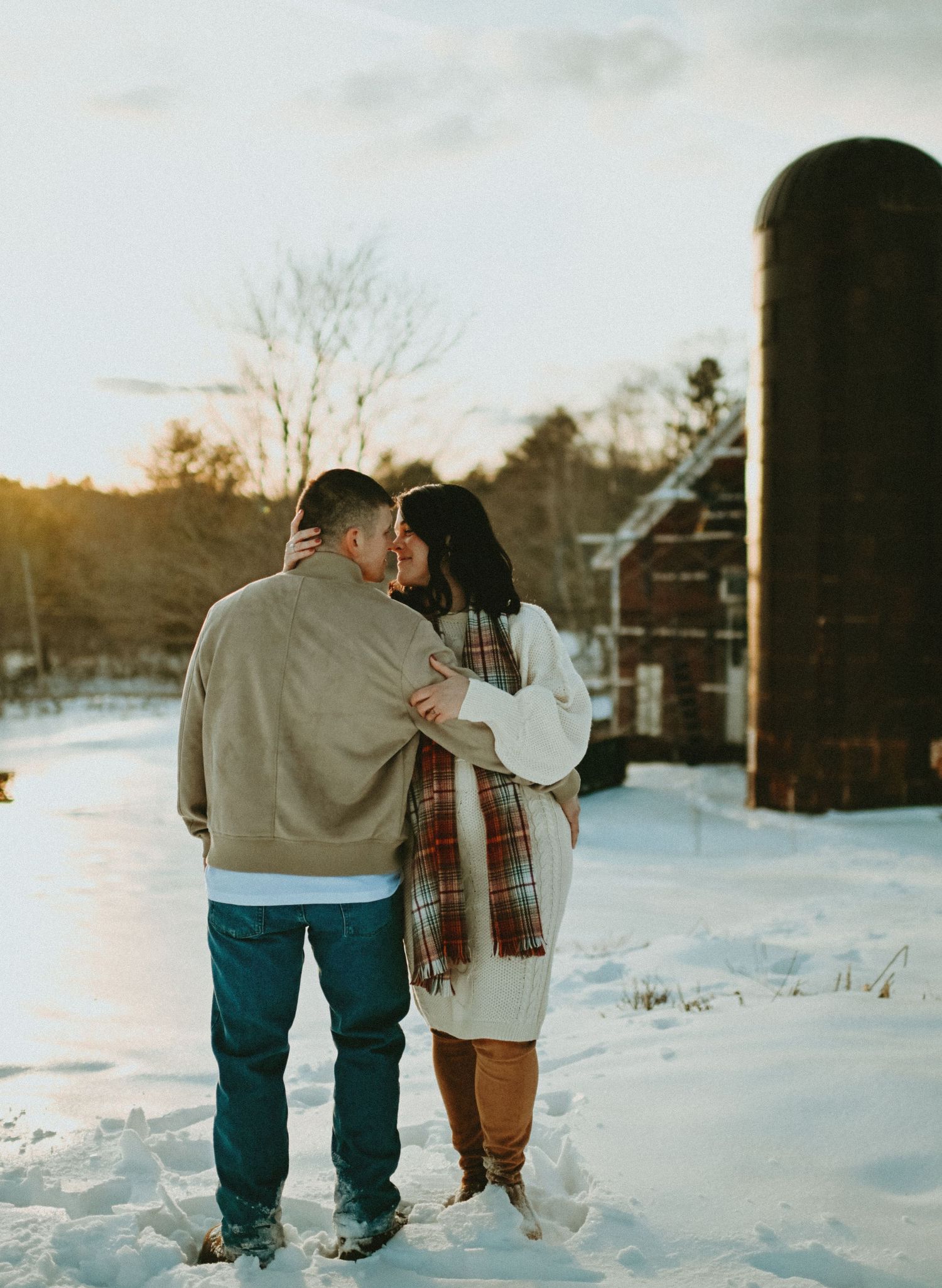 Couple embracing and kissing near a rustic barn silo in a beautiful snowy winter sunset scene.