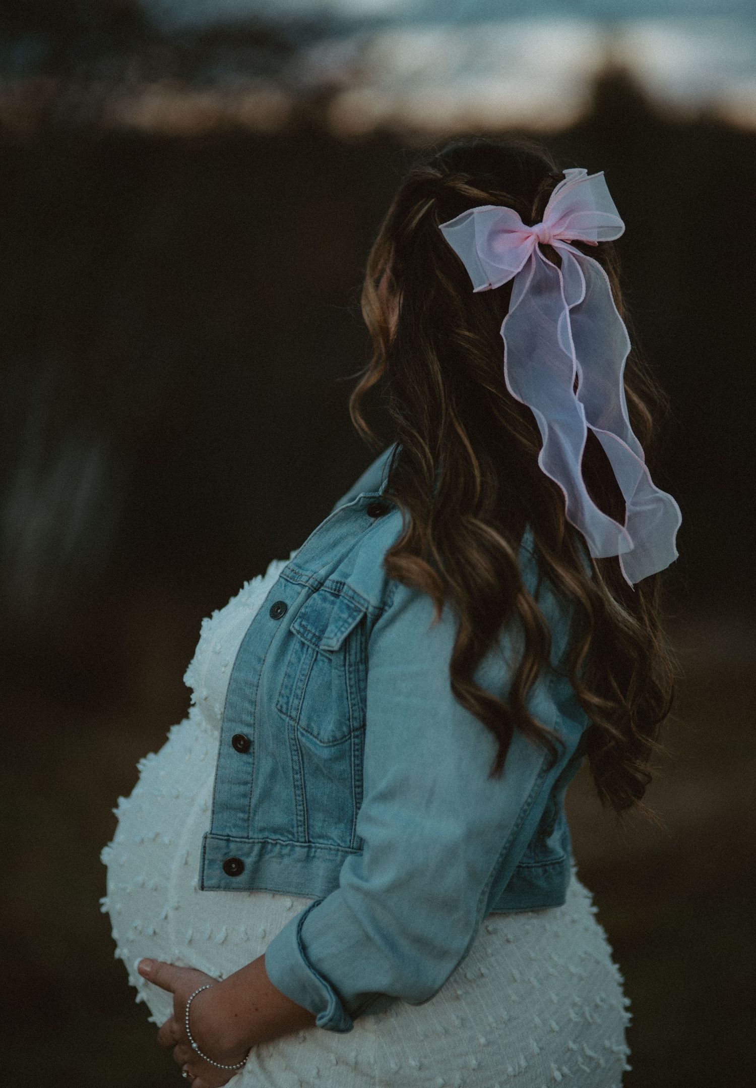 Back view of pregnant woman in denim jacket with pink bow hair accessory.