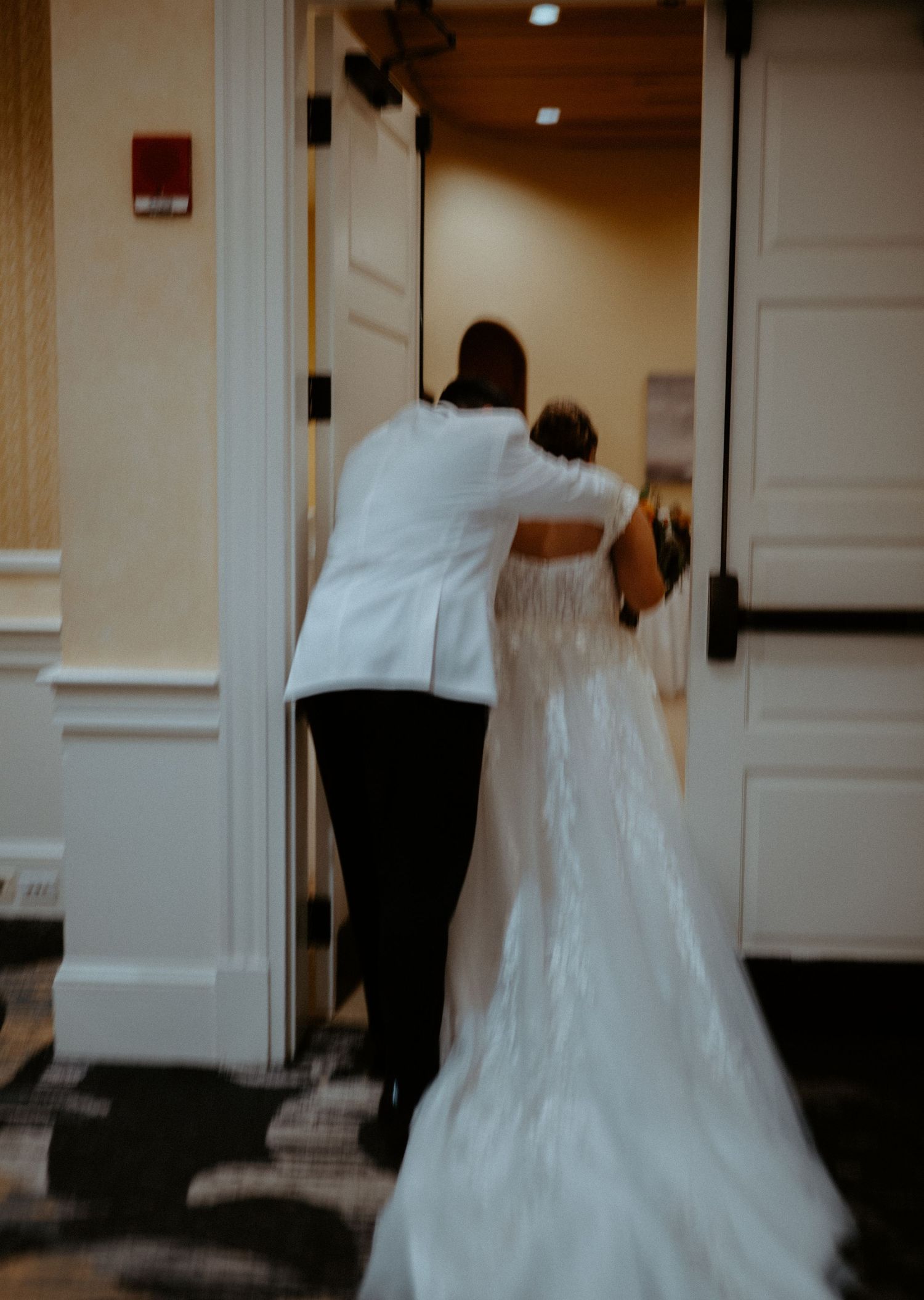 Brides in wedding attire walk through elegant venue doors, One bride in white jacket holding door for bride in lace gown.