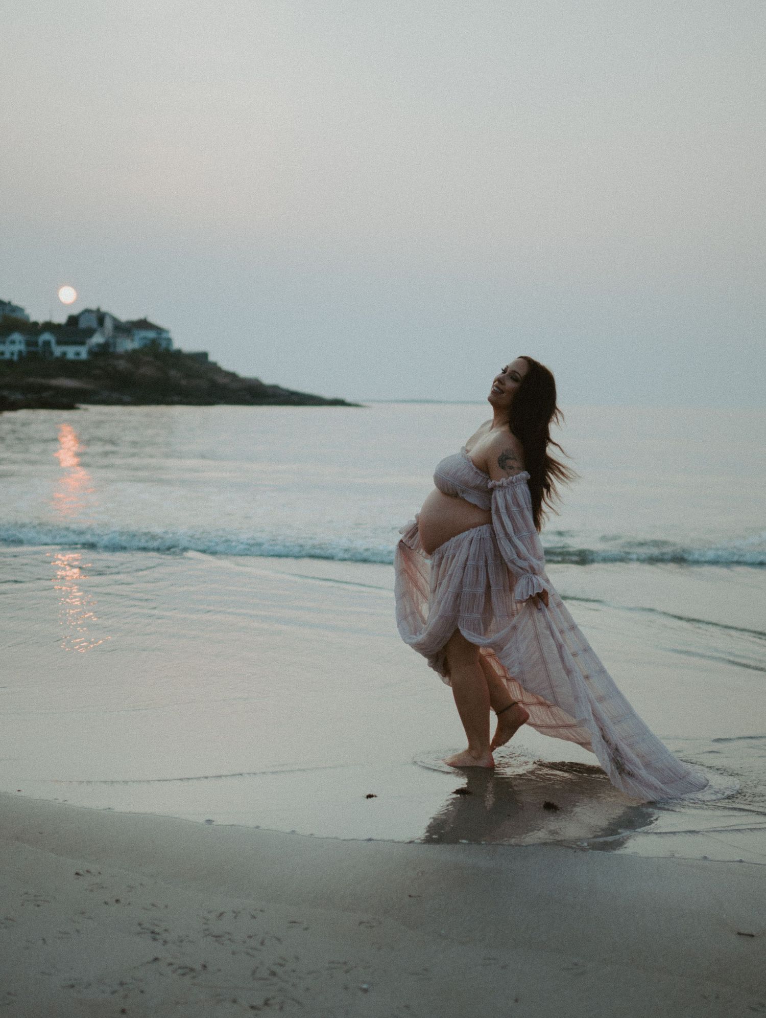 Pregnant woman in flowing pink dress posing gracefully on a beach at sunrise.