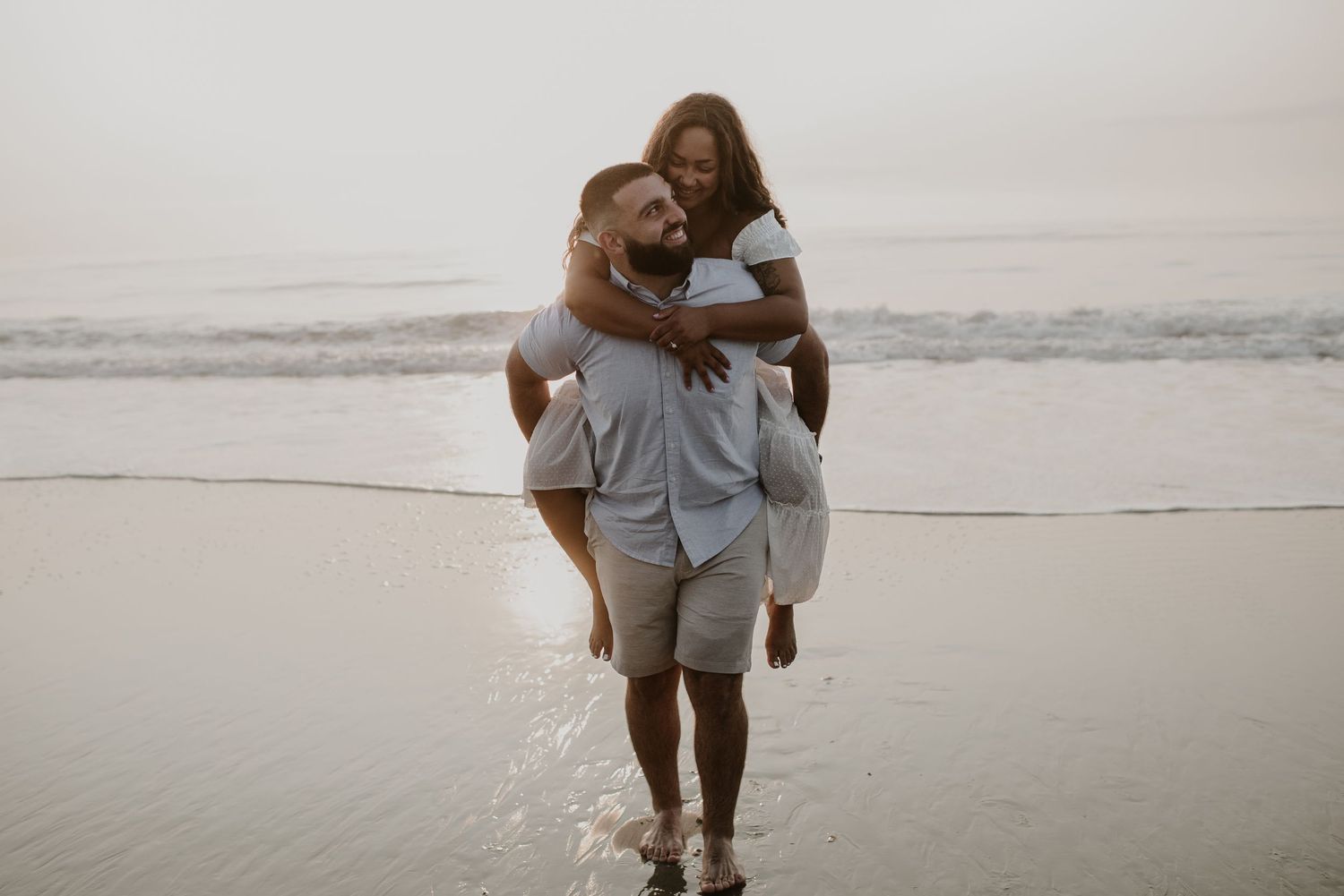 Happy couple enjoying a piggyback ride along the shoreline with gentle ocean waves in background.