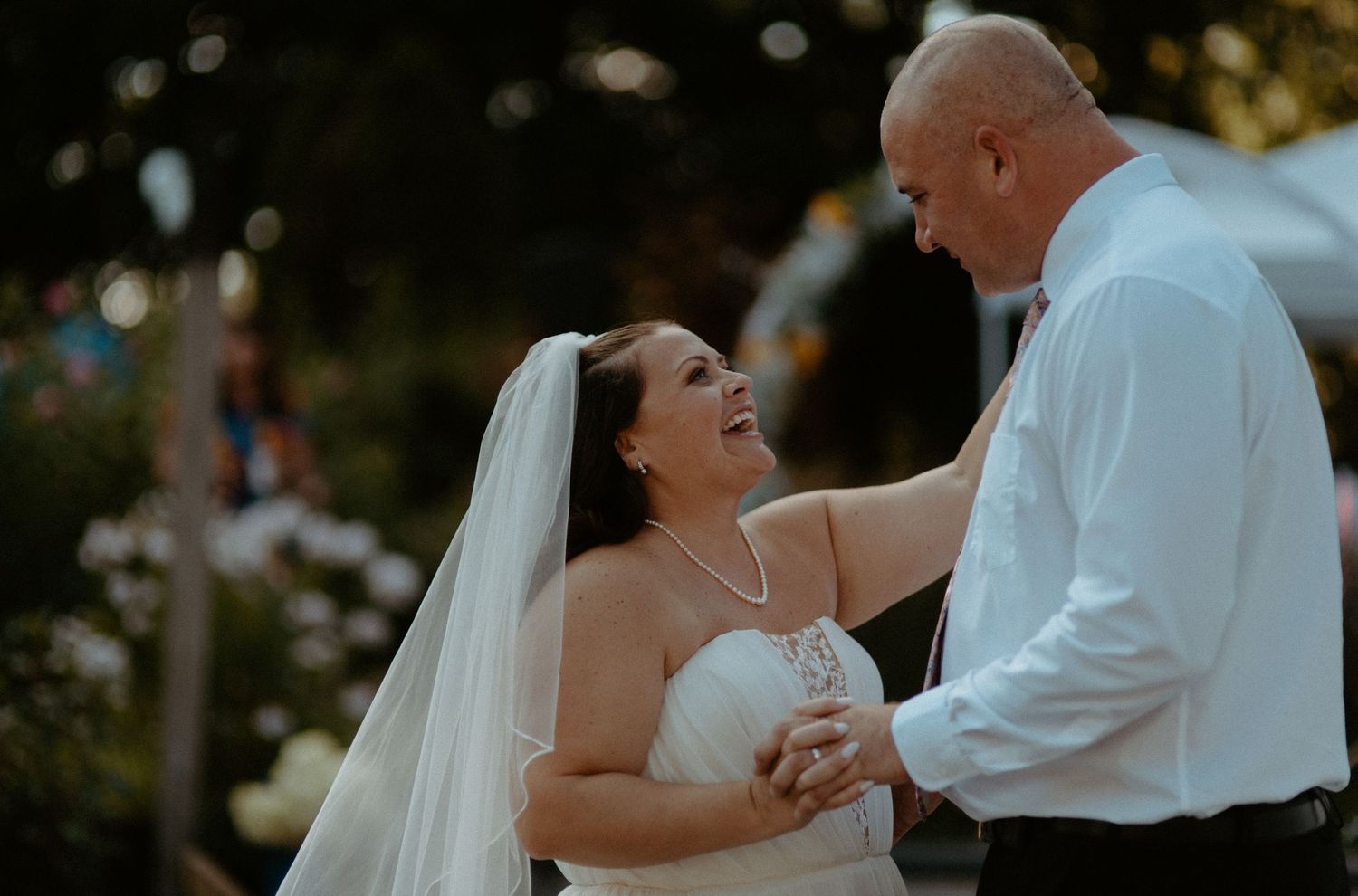 Joyful bride in strapless wedding gown and veil laughing with groom during first dance at outdoor reception.