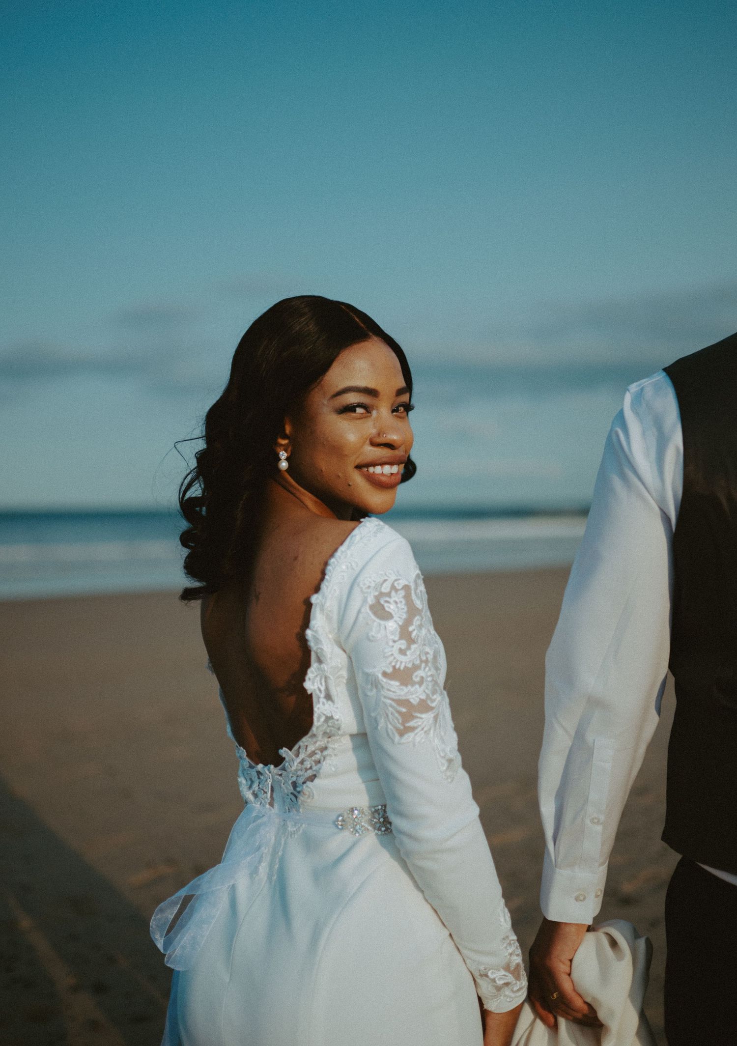 Bride in backless lace wedding gown smiling over shoulder on beach at sunset.