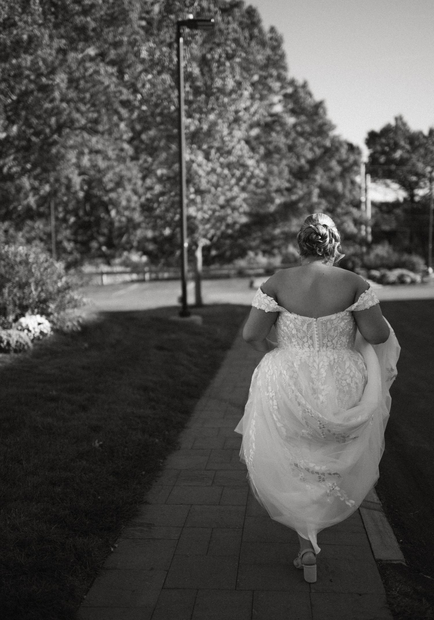 Black and white photo of bride in lace off-shoulder gown walking along a tree-lined pathway.