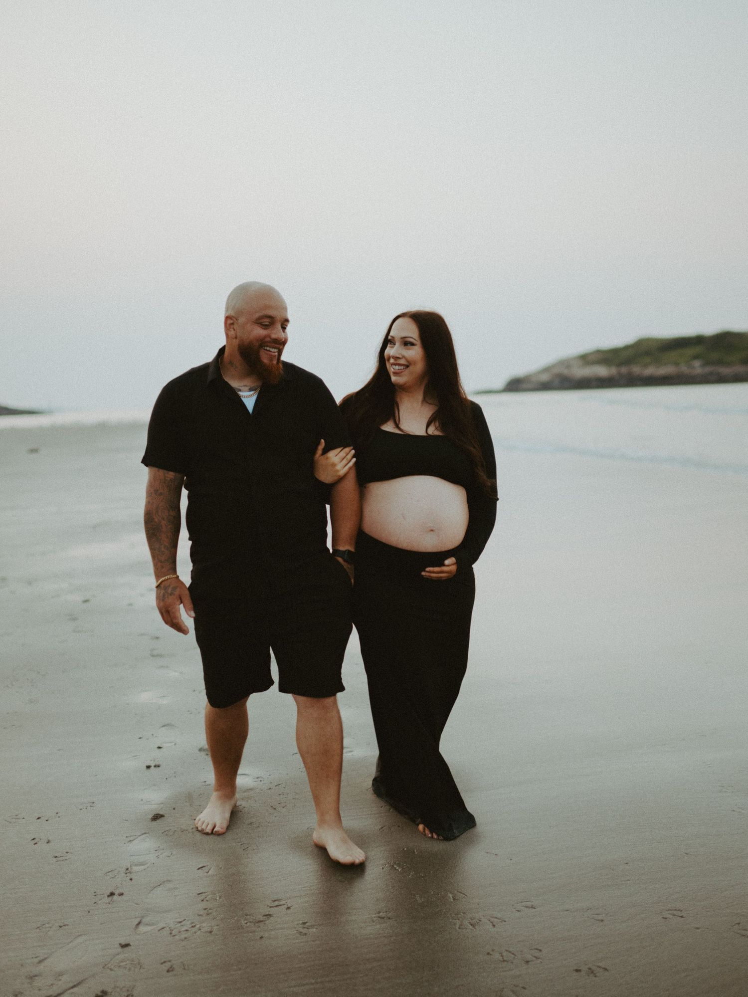 Smiling couple walking on beach, pregnant woman in black crop top and skirt with partner.