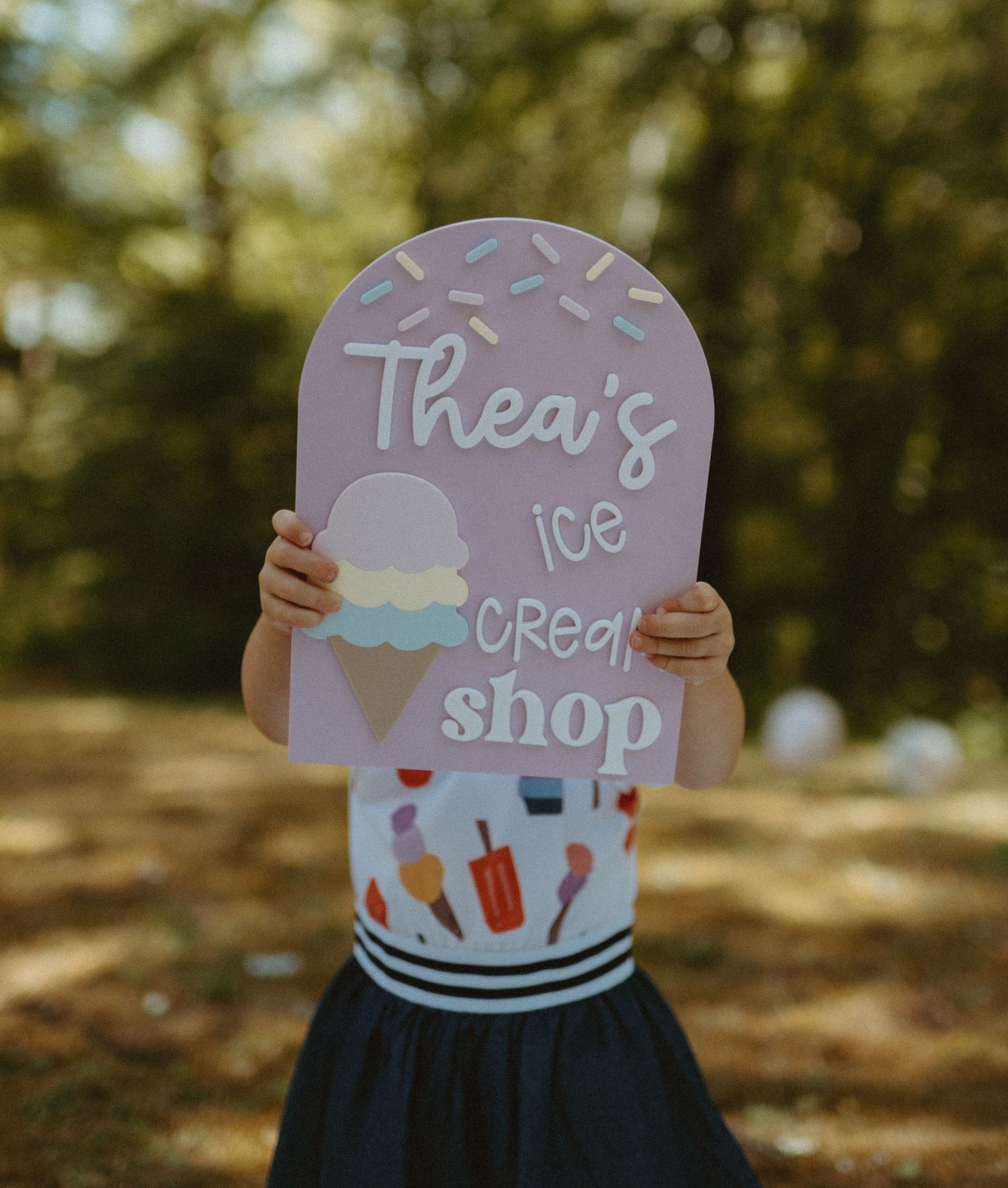 Child holding Ice Cream Shop birthday sign in sunny outdoor setting.
