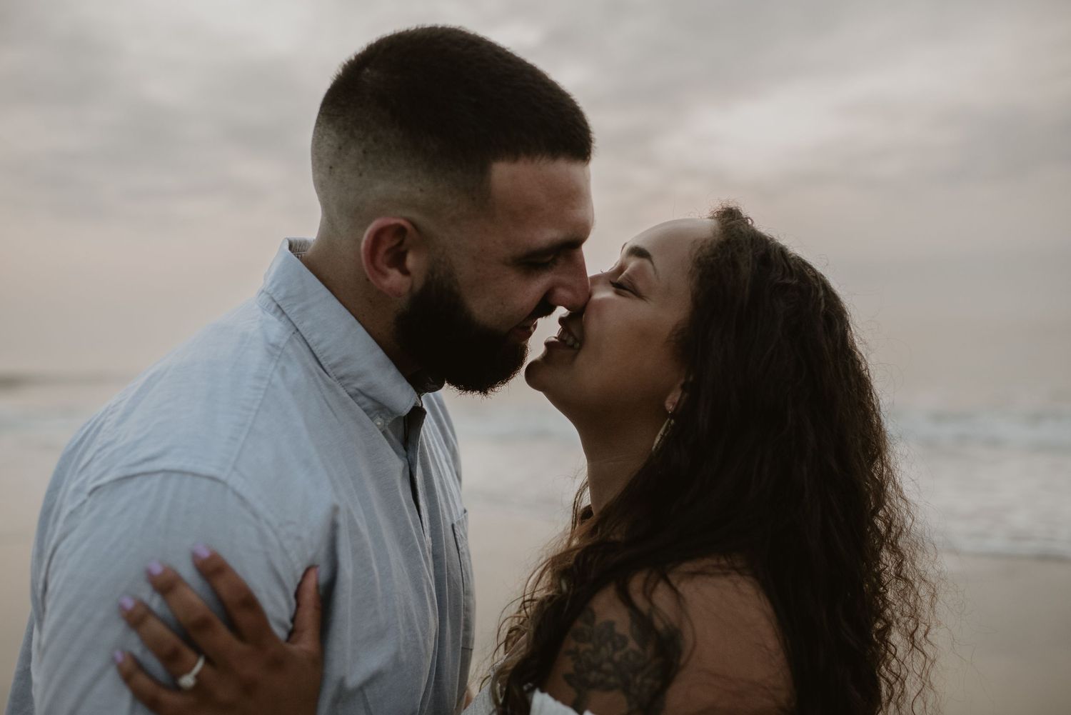 Couple sharing a romantic almost-kiss on the beach at sunset, woman wearing engagement ring.