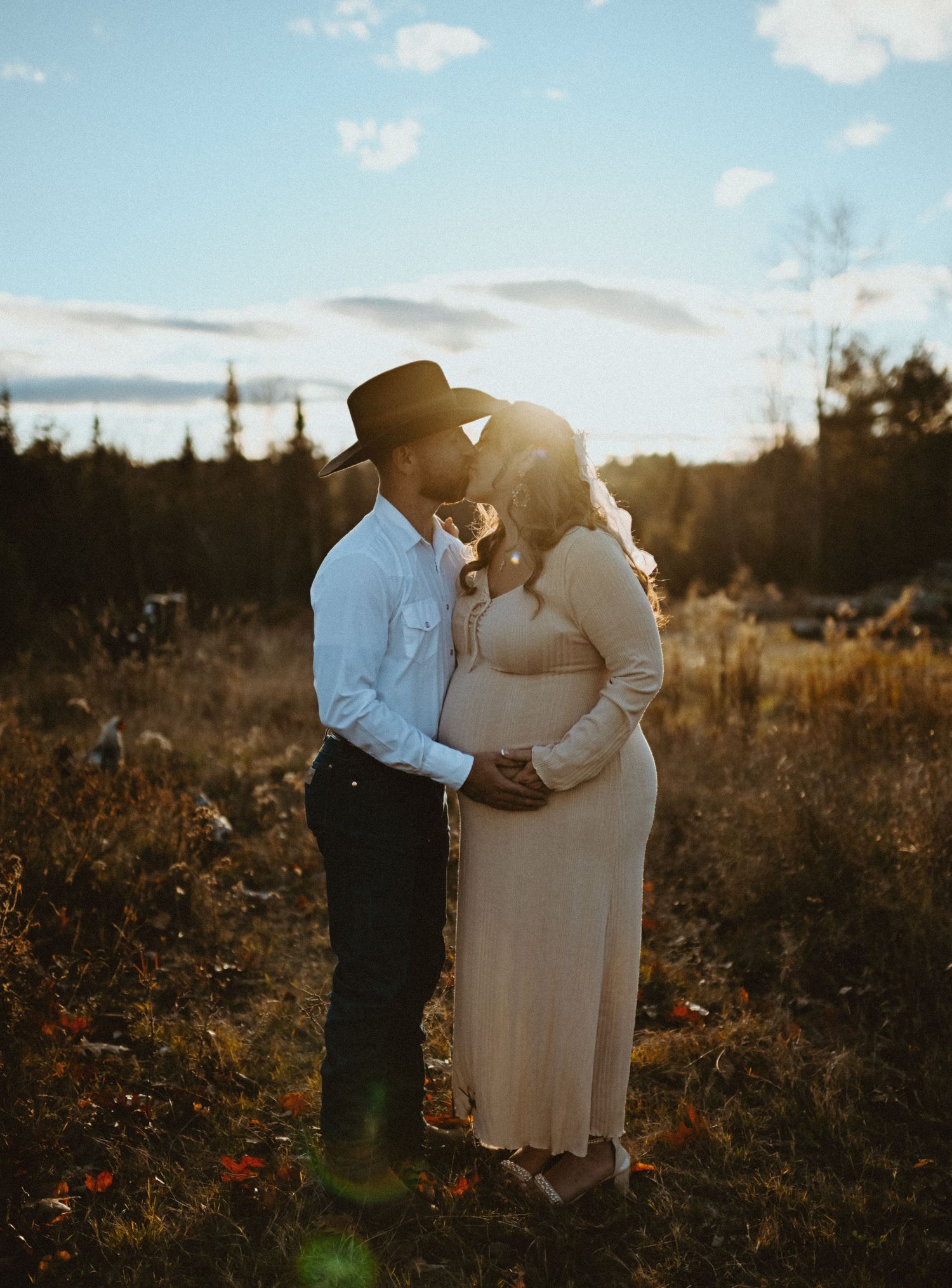 Couple kissing at sunset during maternity photos, man in cowboy hat holds pregnant woman in beige dress.