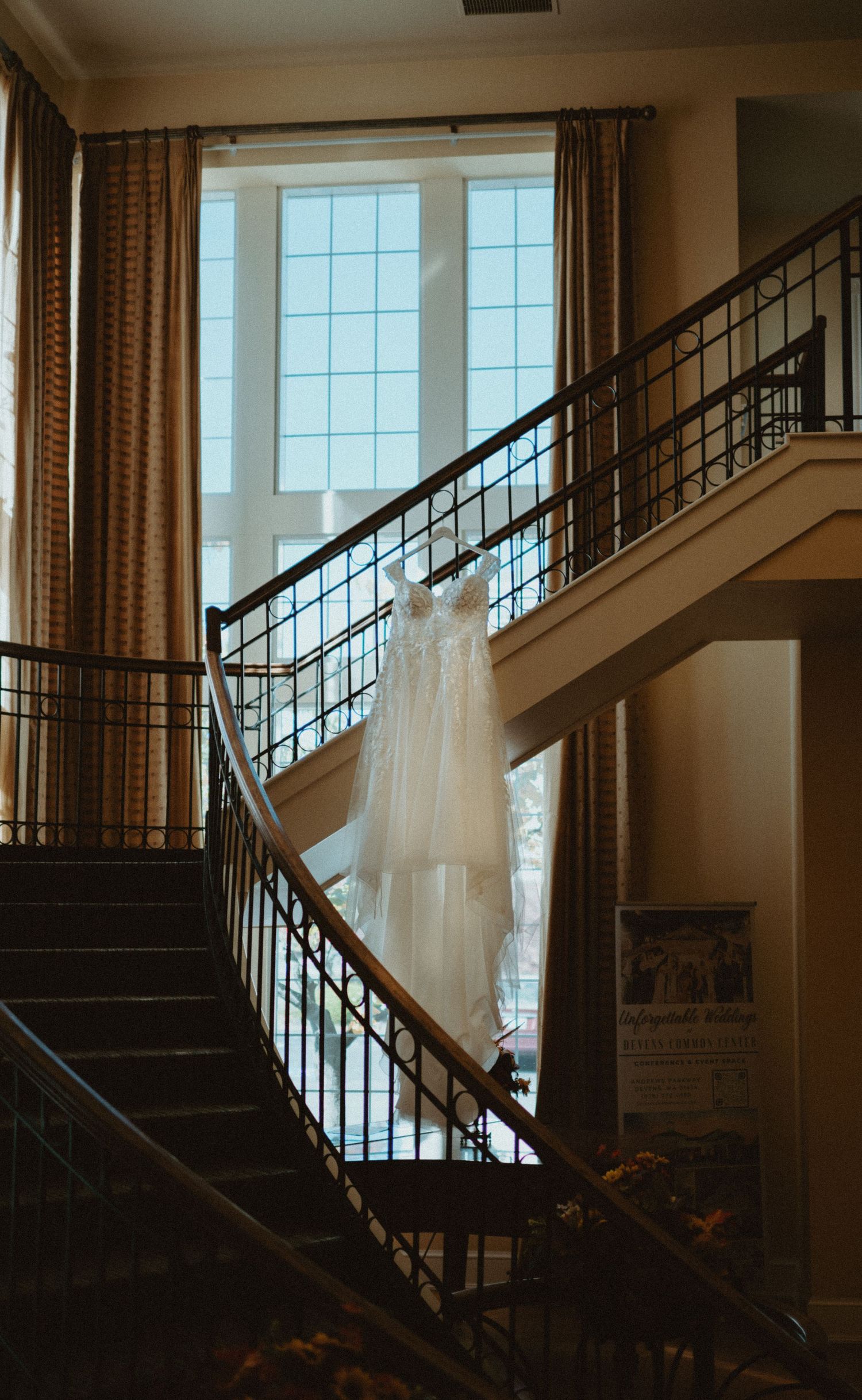 Elegant white lace wedding dress hanging on staircase railing near large window in warm light.