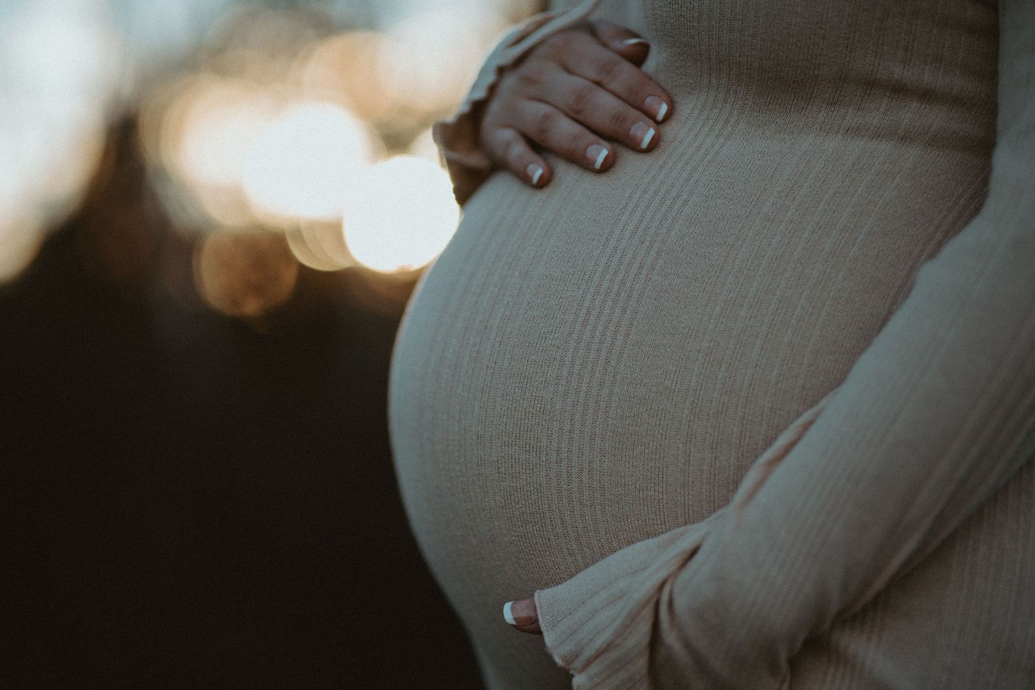 Close-up of pregnant womans baby bump in a cream ribbed maternity dress with soft bokeh background.