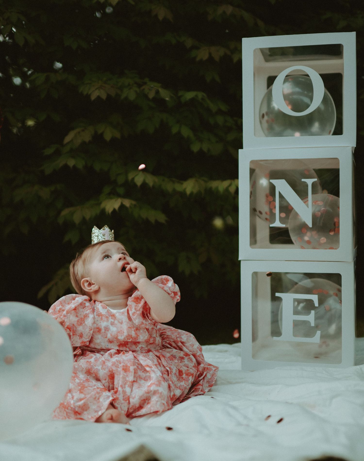 Baby girl in pink dress sitting beside stacked ONE letter blocks for first birthday.