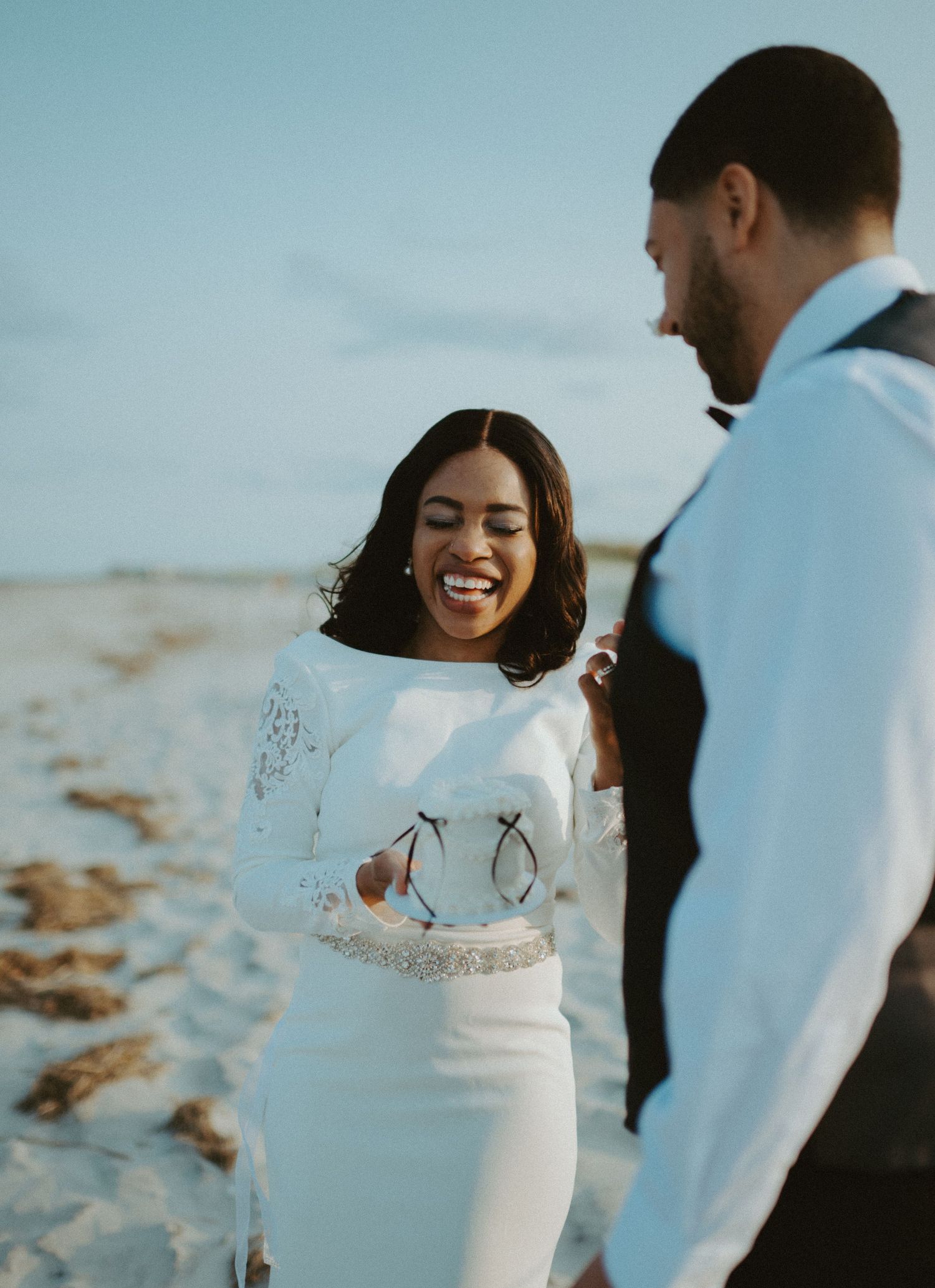 Joyful bride in long-sleeve lace wedding gown laughs with groom on sunny beach wedding day.