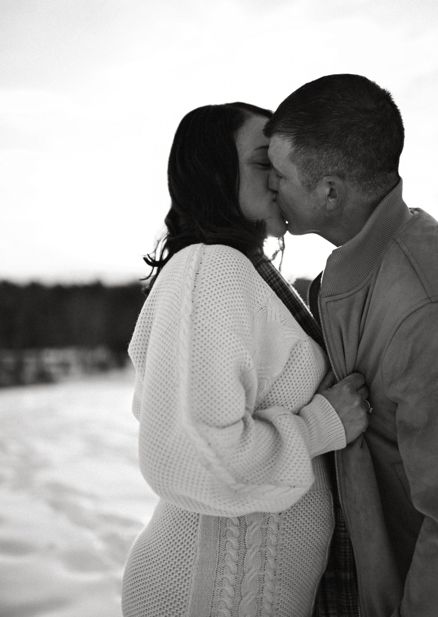 Black and white photo of a couple kissing in a snowy outdoor setting, woman in cozy knit sweater.