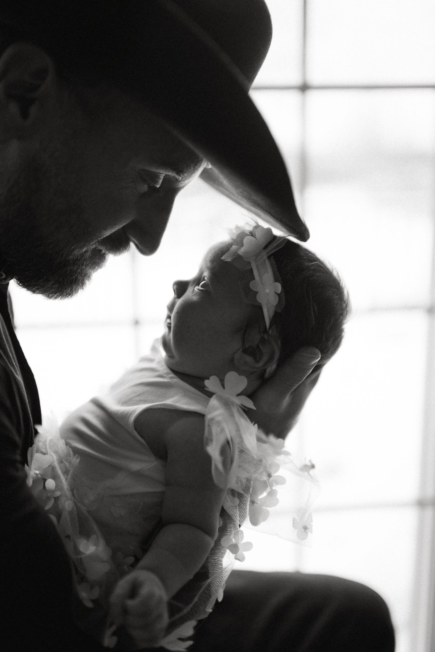 Black and white portrait of a bearded father in cowboy hat gazing lovingly at his newborn baby.