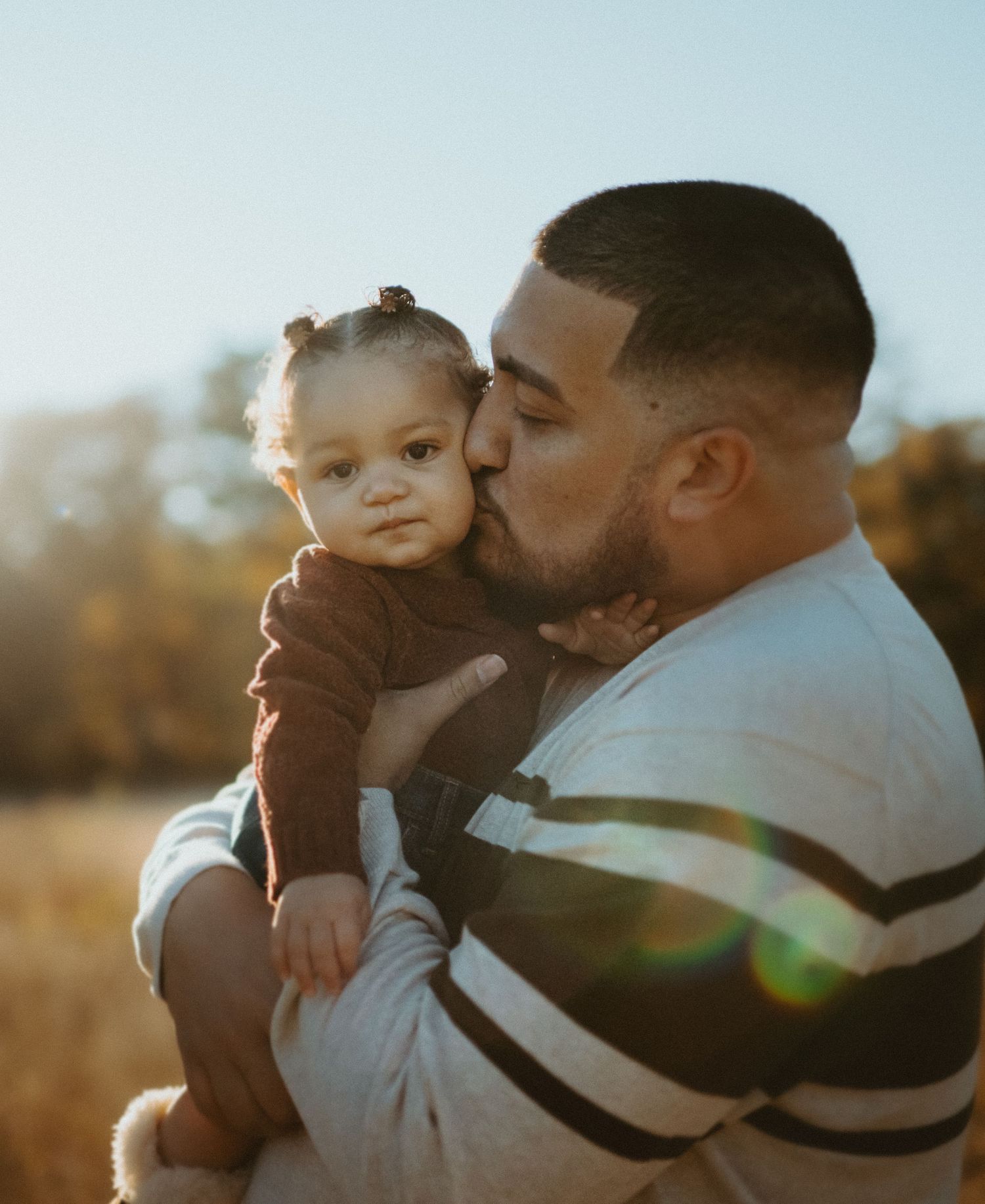 Father kissing his baby girl on cheek in sunny autumn field outdoors.