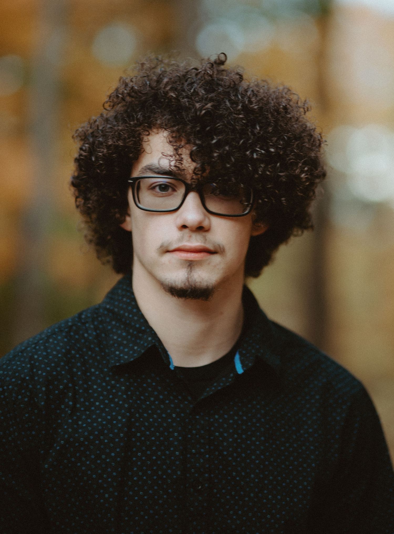 Young man with curly hair and glasses wearing black shirt in fall outdoor portrait.