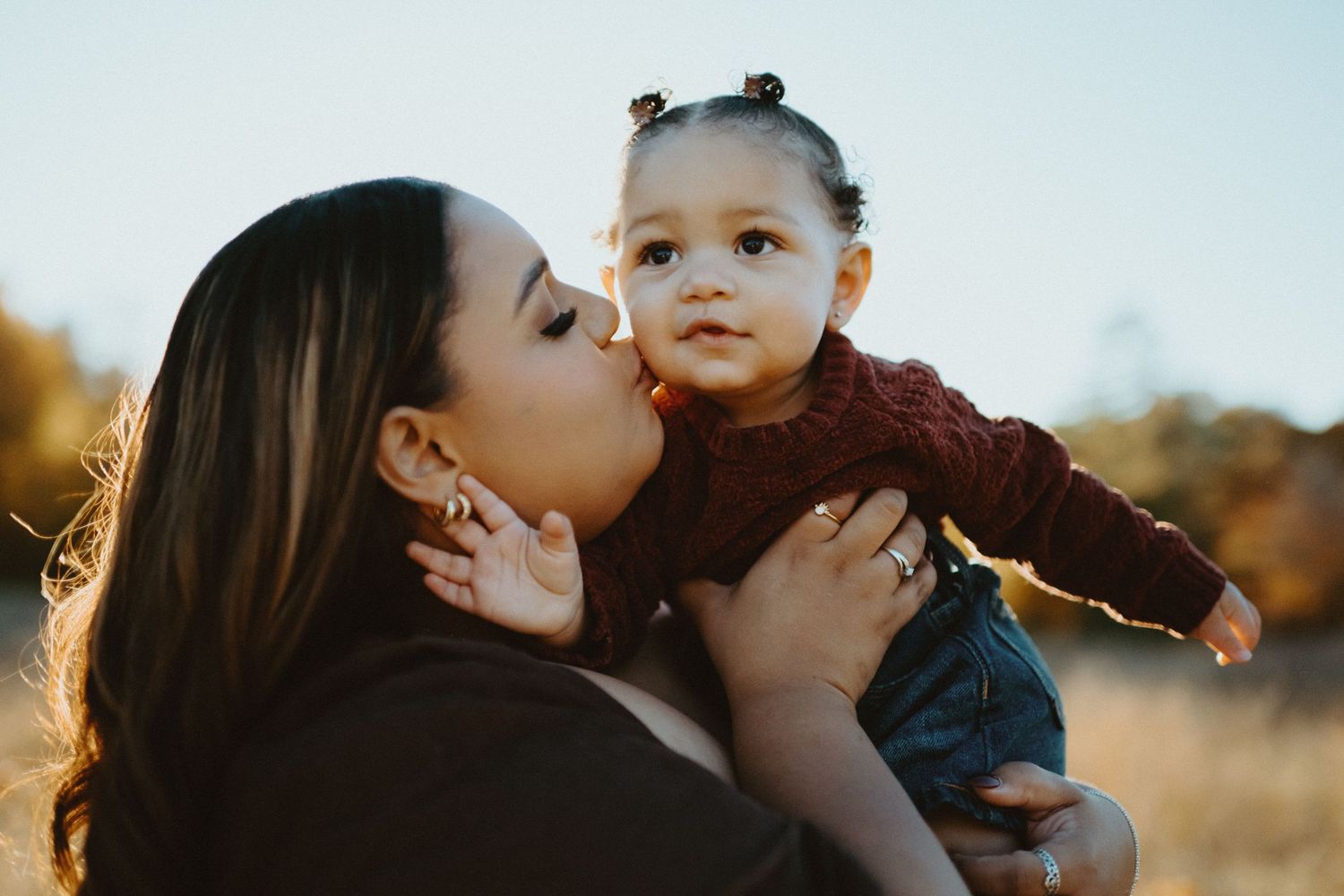 Mother kissing her toddler daughter on cheek in warm golden sunlight outdoors.