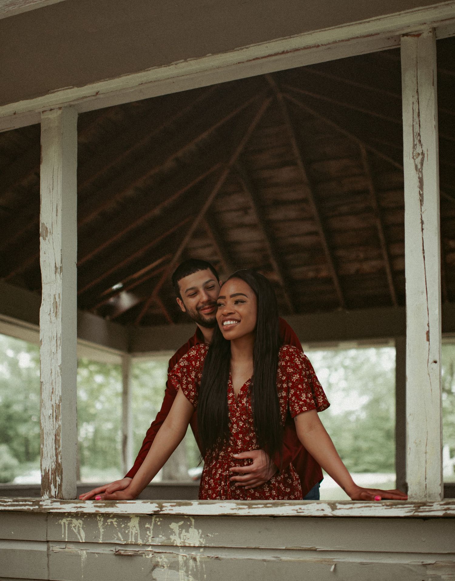 Smiling couple posing in a weathered white gazebo window, woman in red floral dress embraced by partner.