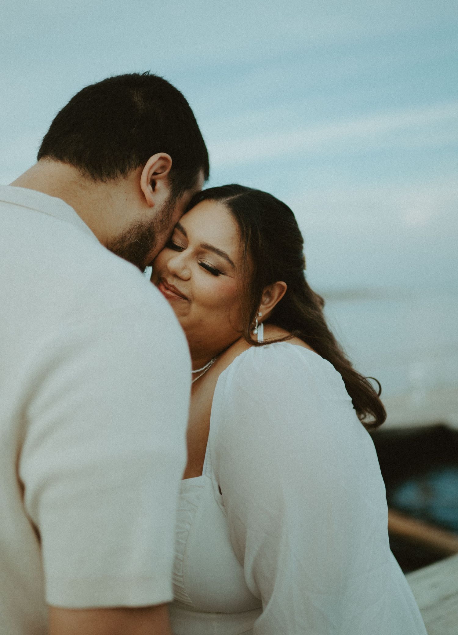 Couple sharing a tender cheek kiss moment outdoors with ocean backdrop at dusk.