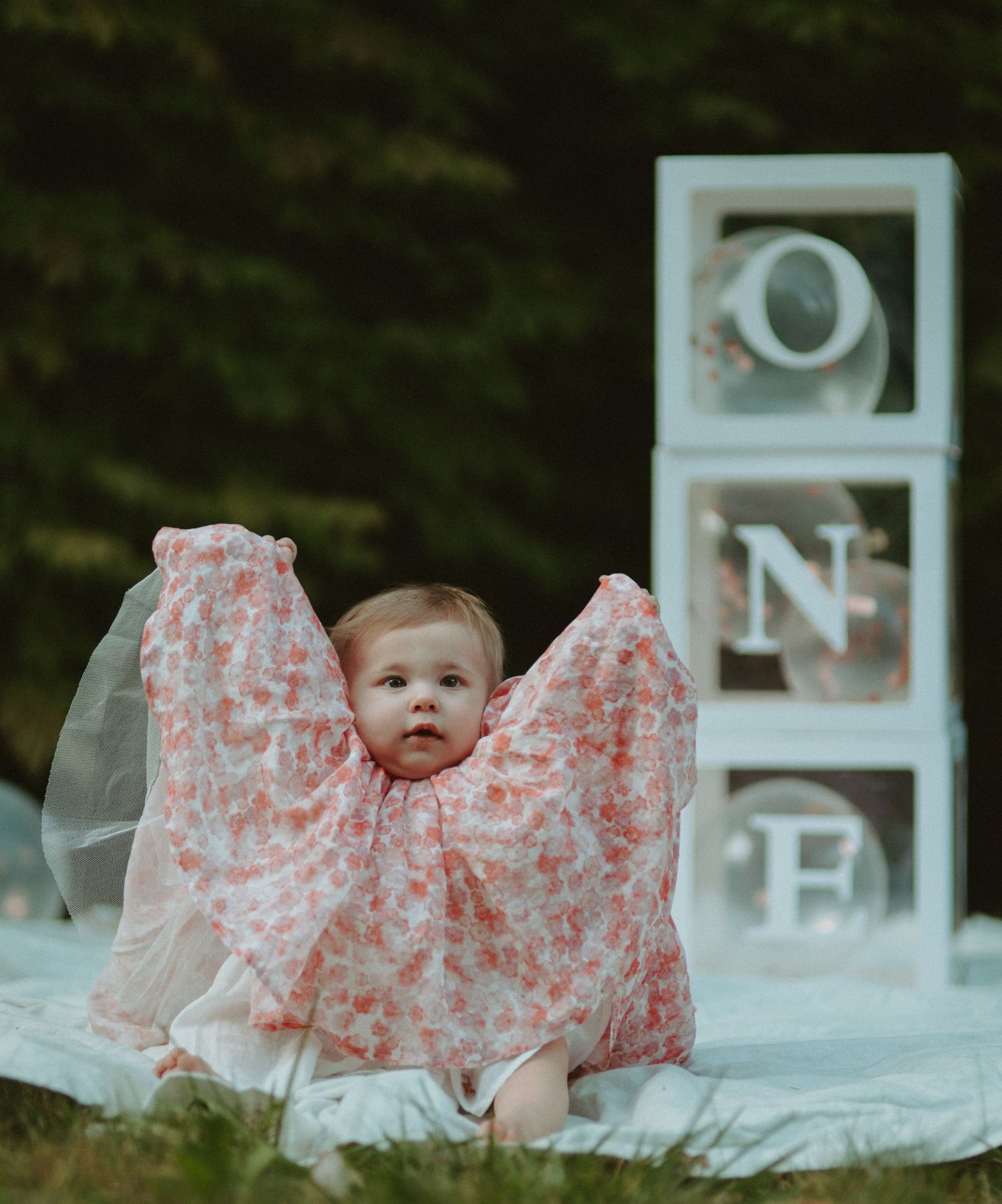 Baby girl in pink floral dress sits before stacked ONE letter blocks for first birthday photos.