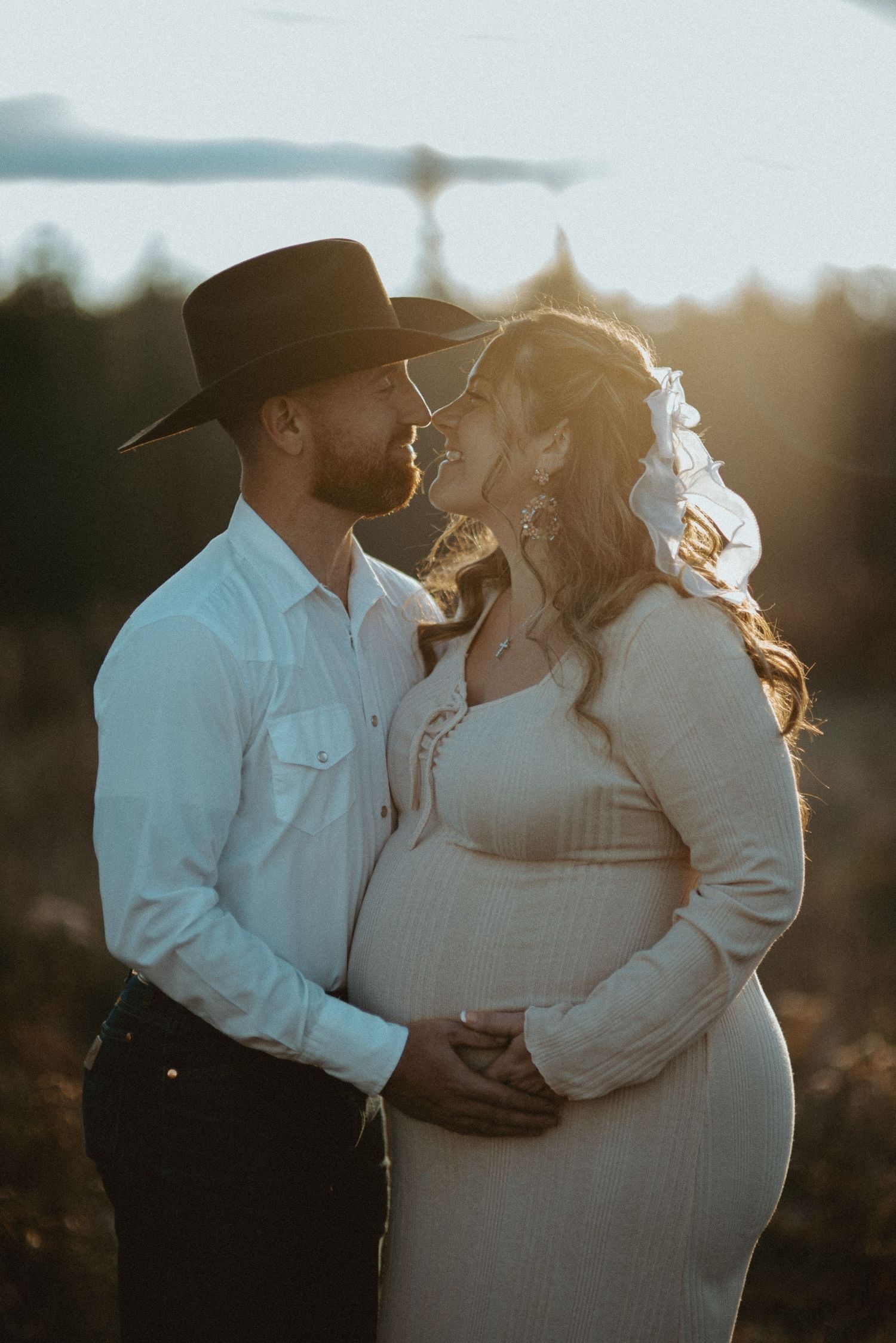 Pregnant couple sharing a romantic moment at golden sunset, man in cowboy hat and white shirt.