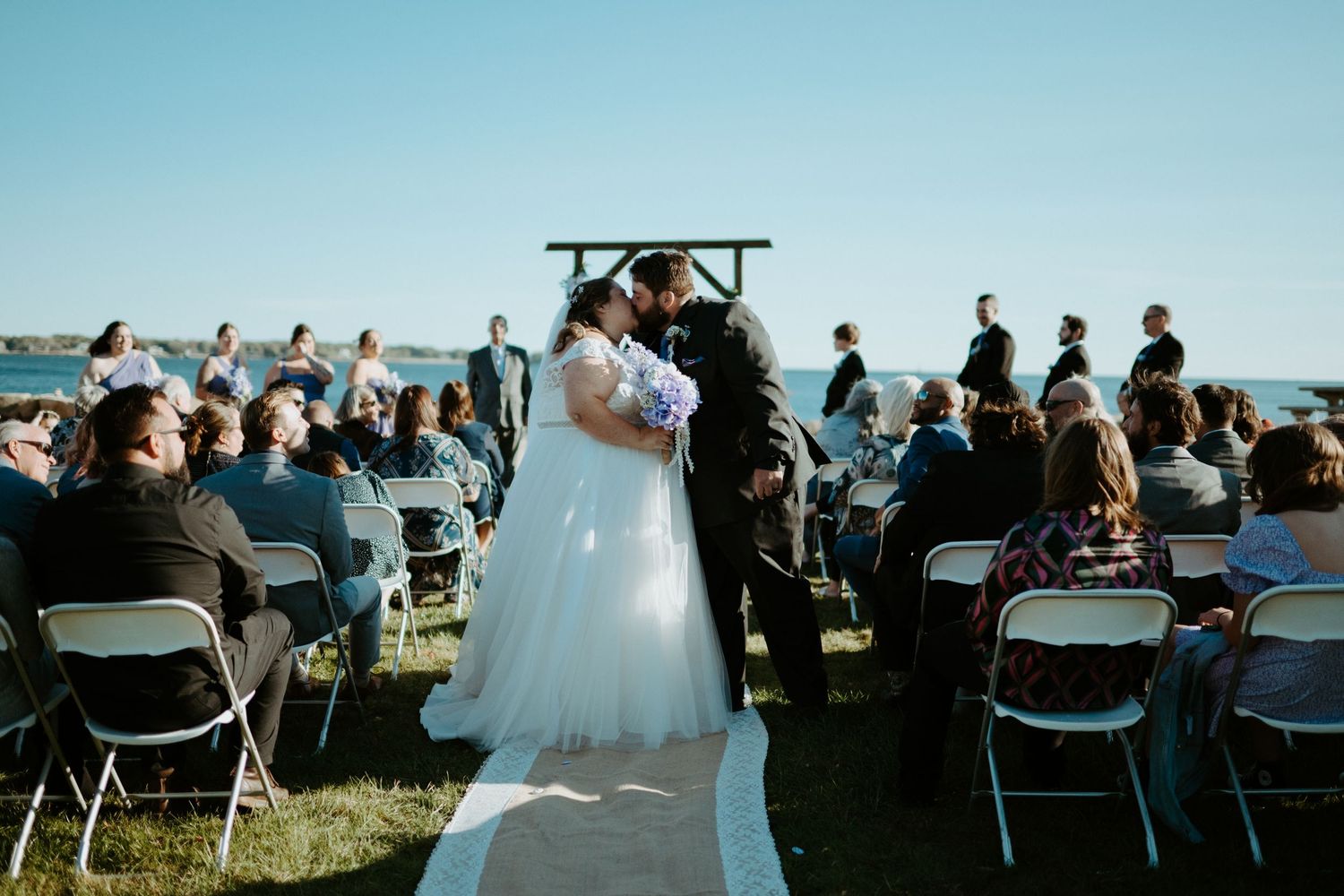 Bride and groom share first kiss at outdoor oceanside wedding ceremony with seated guests.
