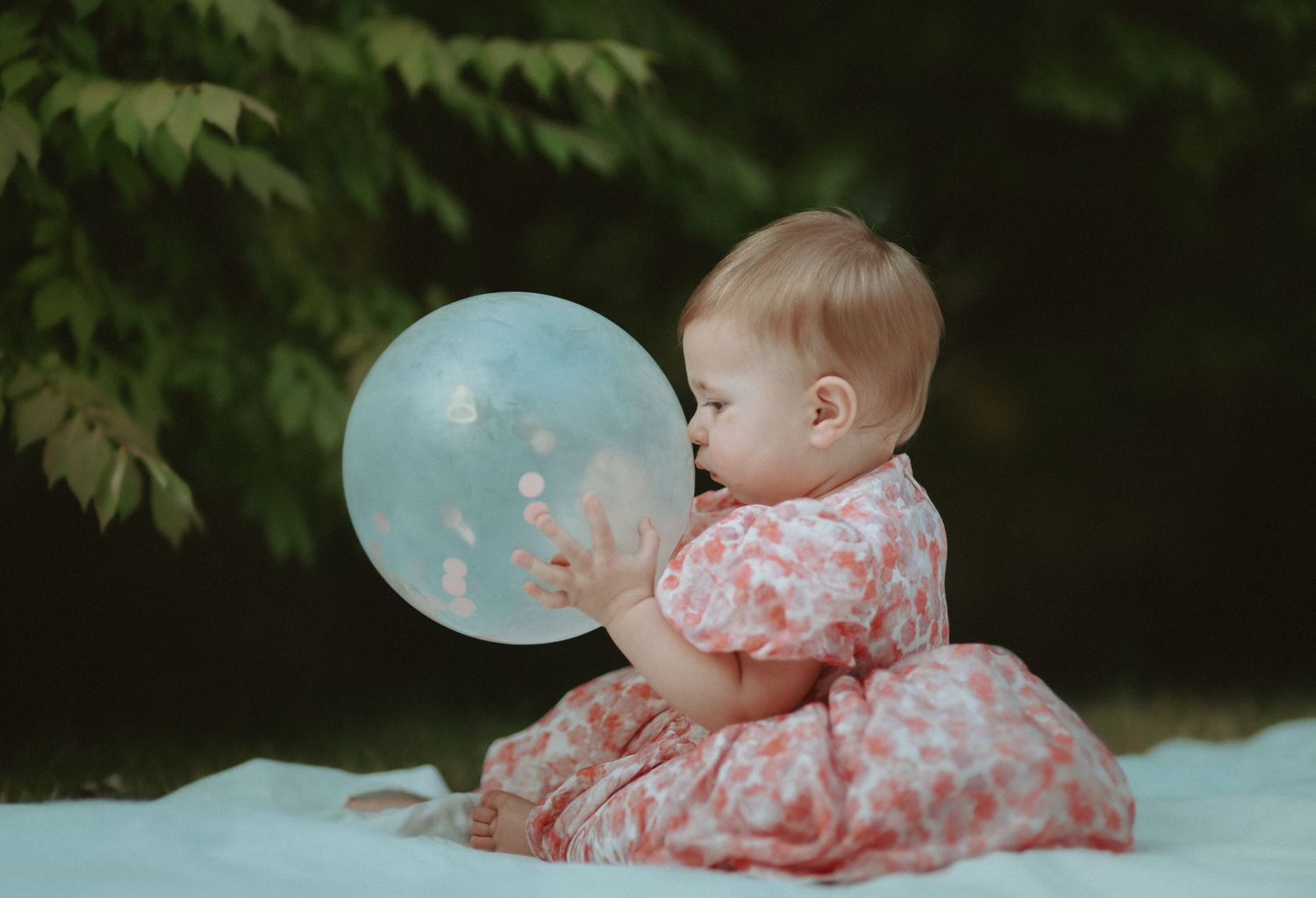 Adorable baby girl in pink floral dress plays with a translucent balloon outdoors.