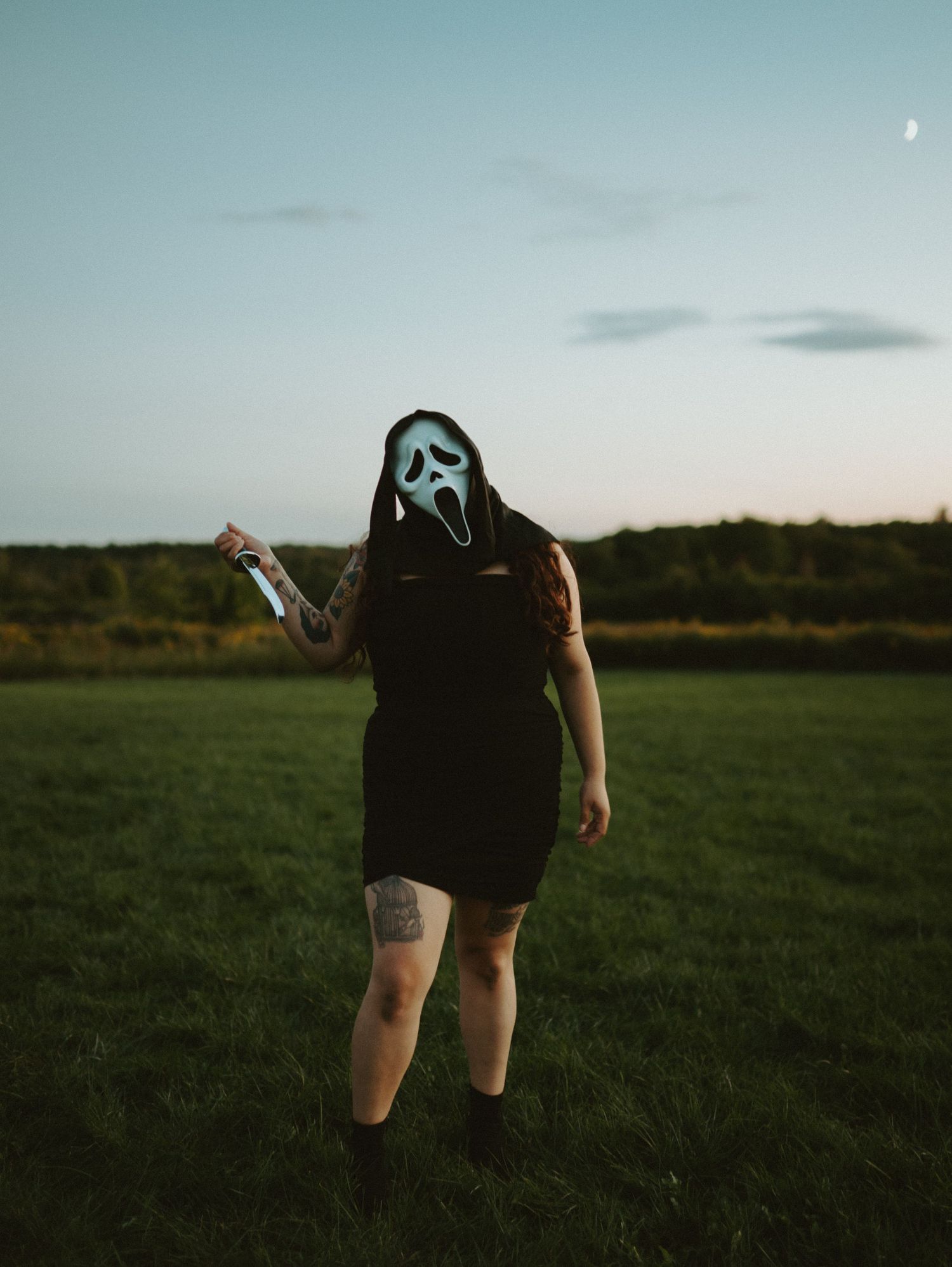 Woman in black dress wearing Ghostface Halloween mask holding a knife in an open field at dusk.