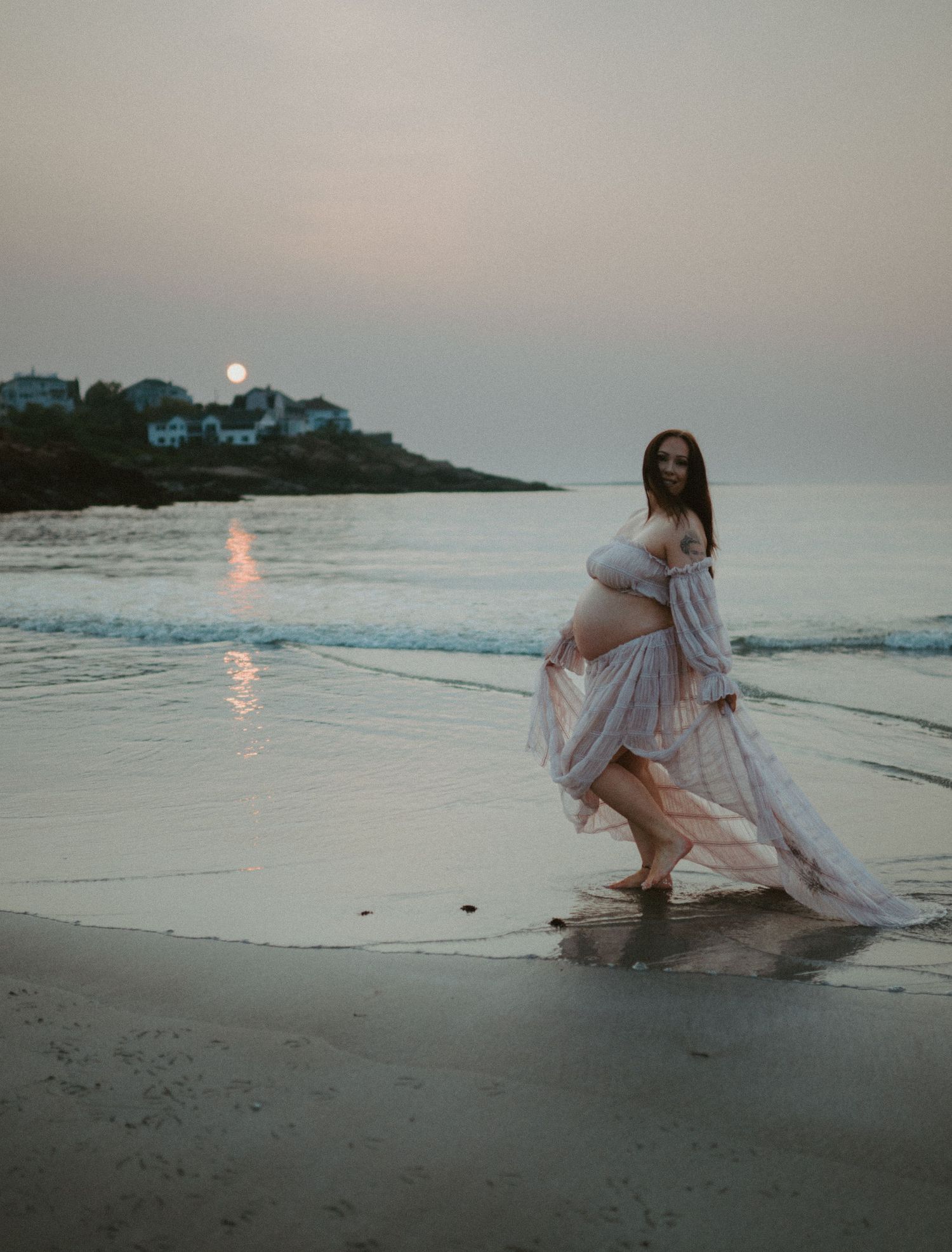 Pregnant woman in pink gown walking on a beach with ocean backdrop. Sunrise in the background.