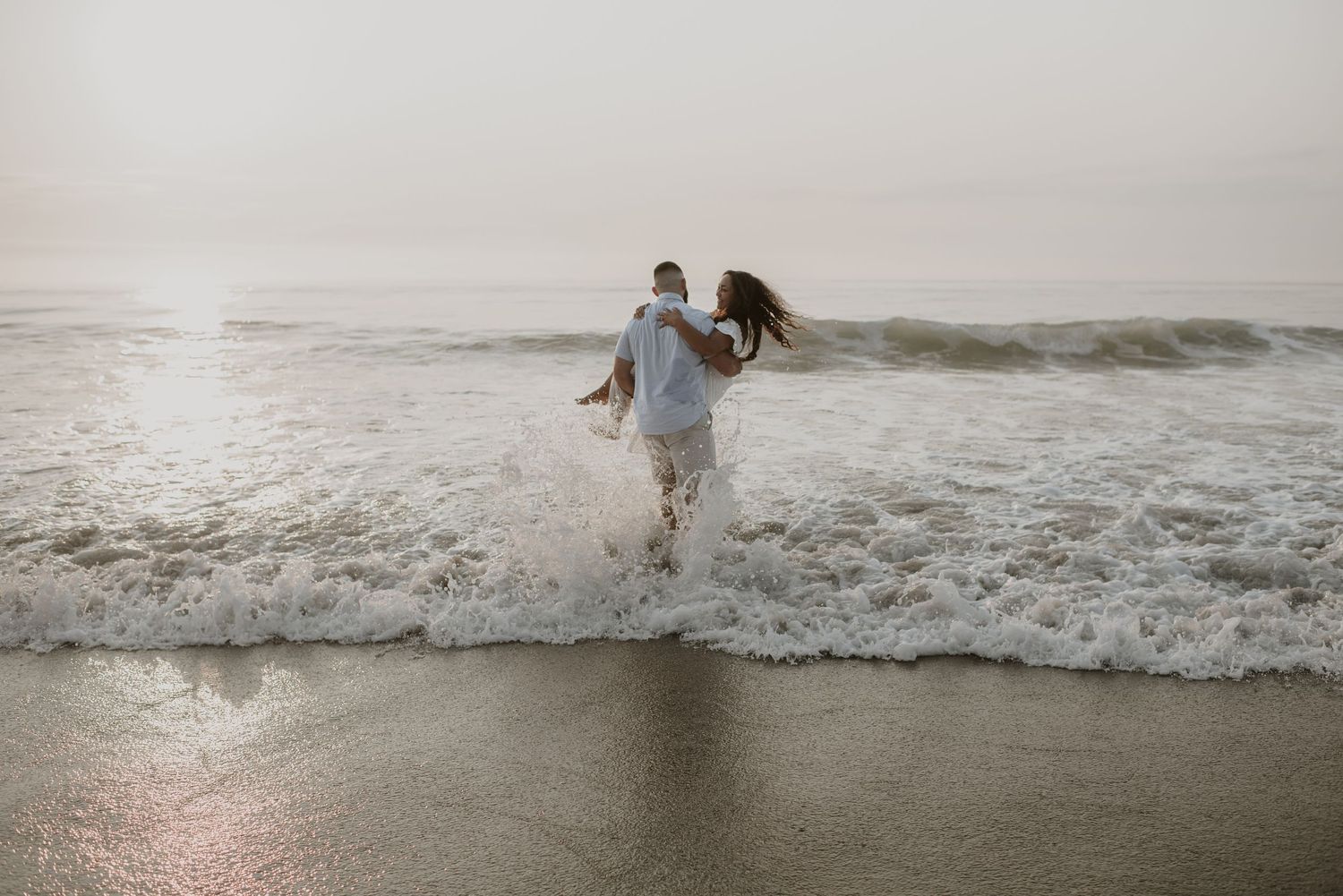 Couple running playfully through ocean waves on the beach, splashing in the surf together.