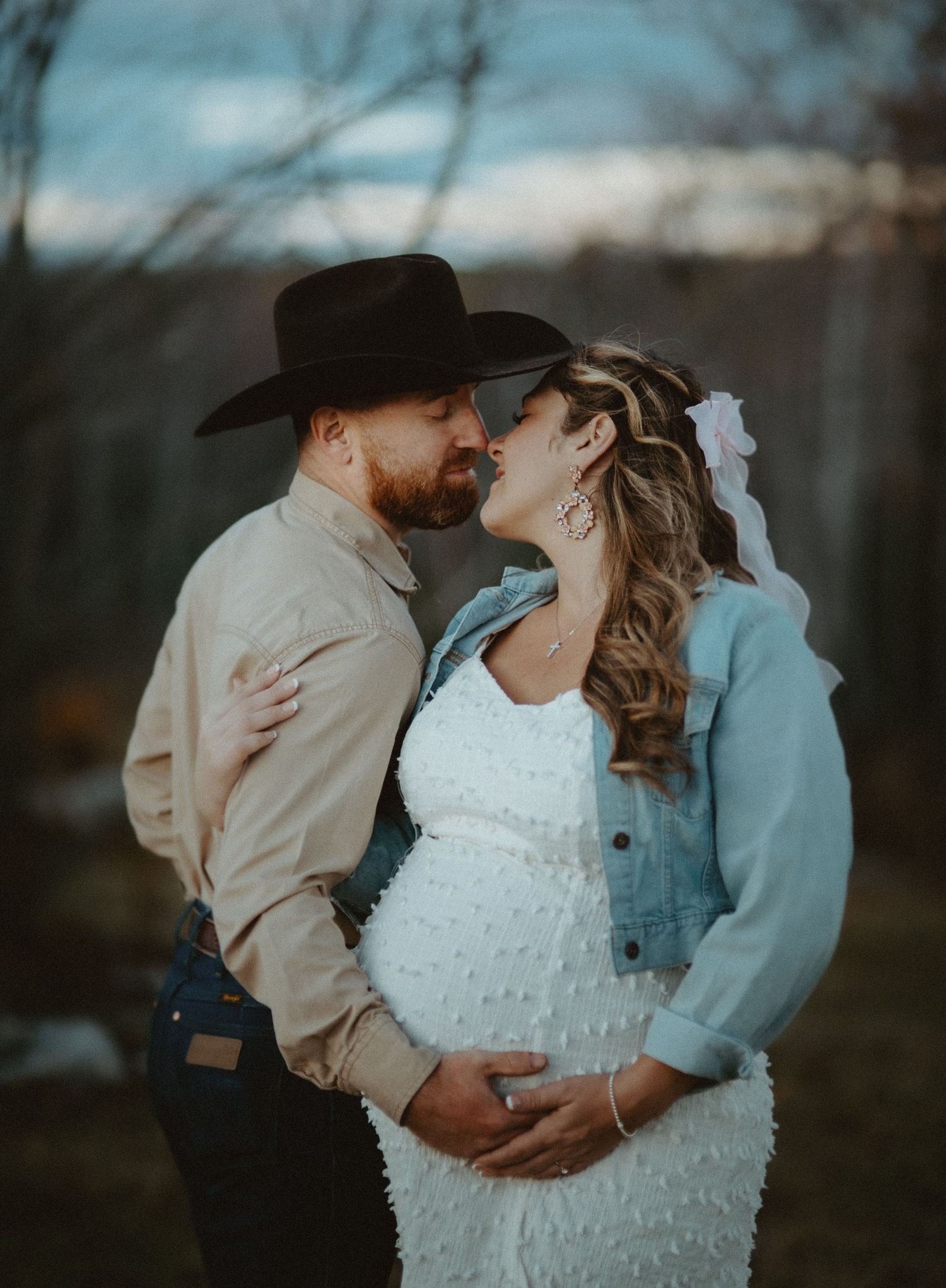 Couple sharing a kiss during maternity shoot, man in cowboy hat embraces pregnant woman in white dress.