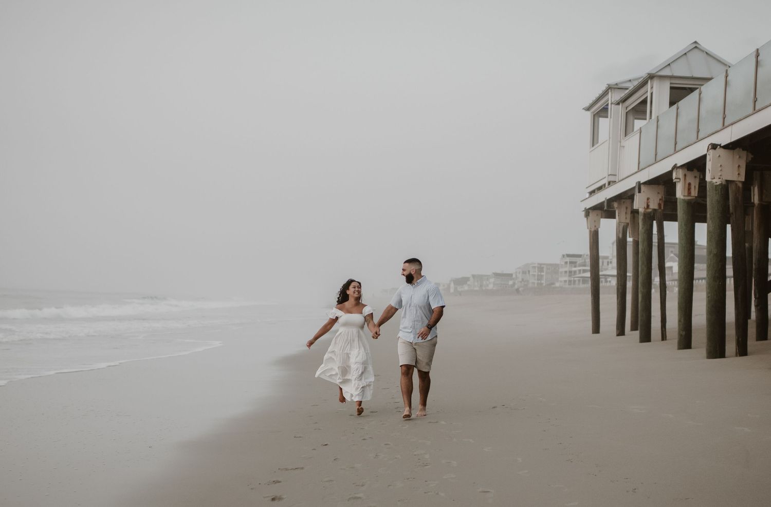 Couple in white outfits walking hand in hand on misty beach near wooden pier.