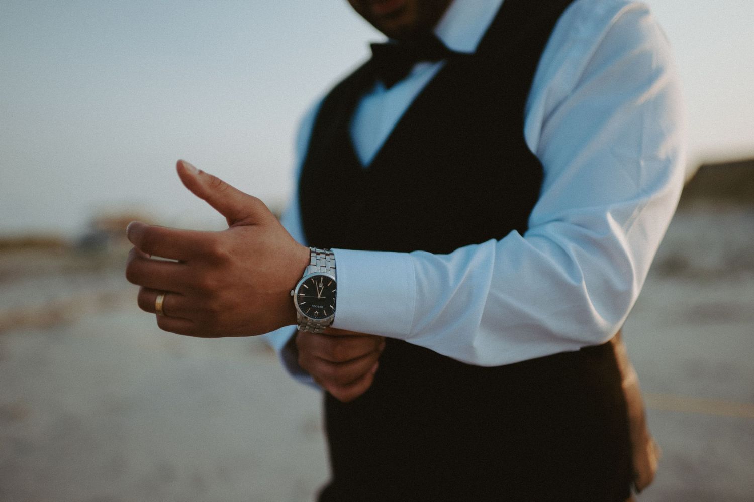 Close-up of groom in black vest and white shirt adjusting silver watch on his wrist at beach.