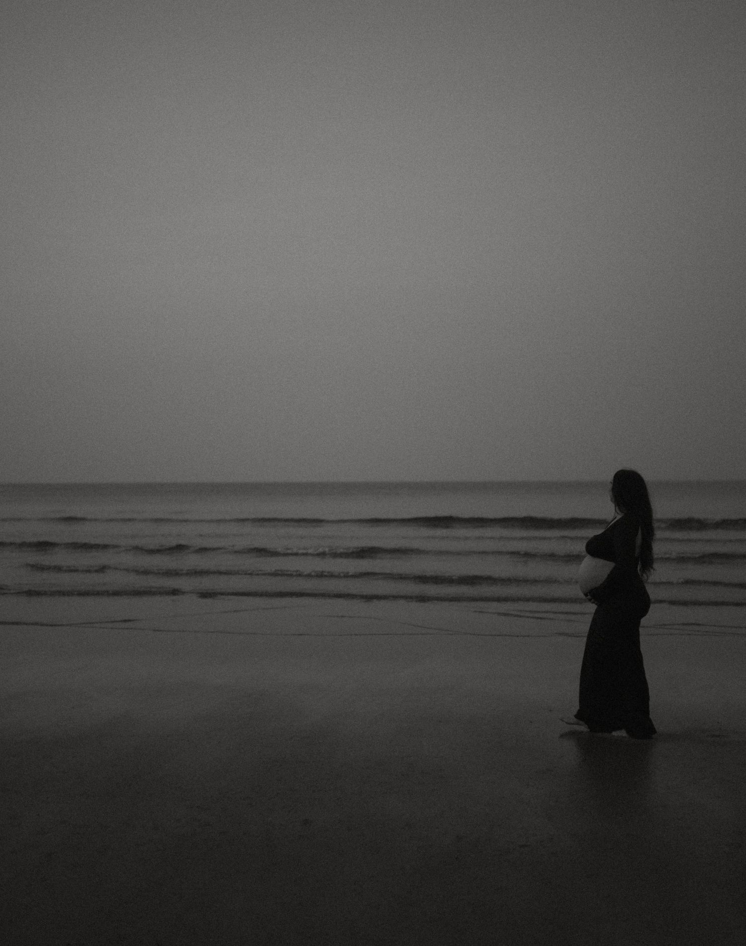 Black and white silhouette of pregnant woman standing on beach facing calm ocean waves.