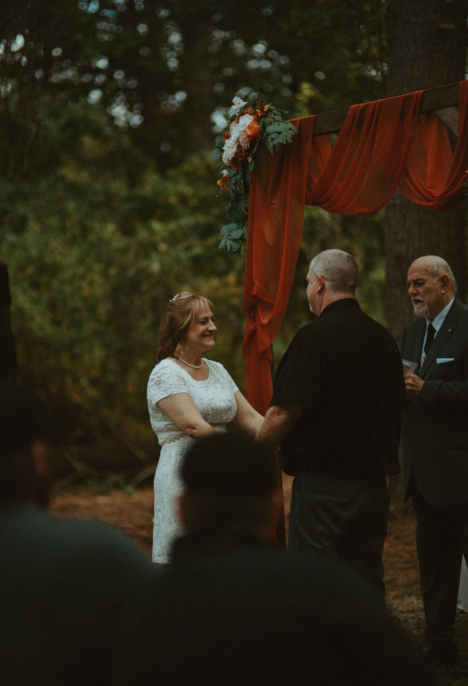 Bride and groom holding hands at outdoor wedding ceremony under rustic orange draped arch in wooded setting.