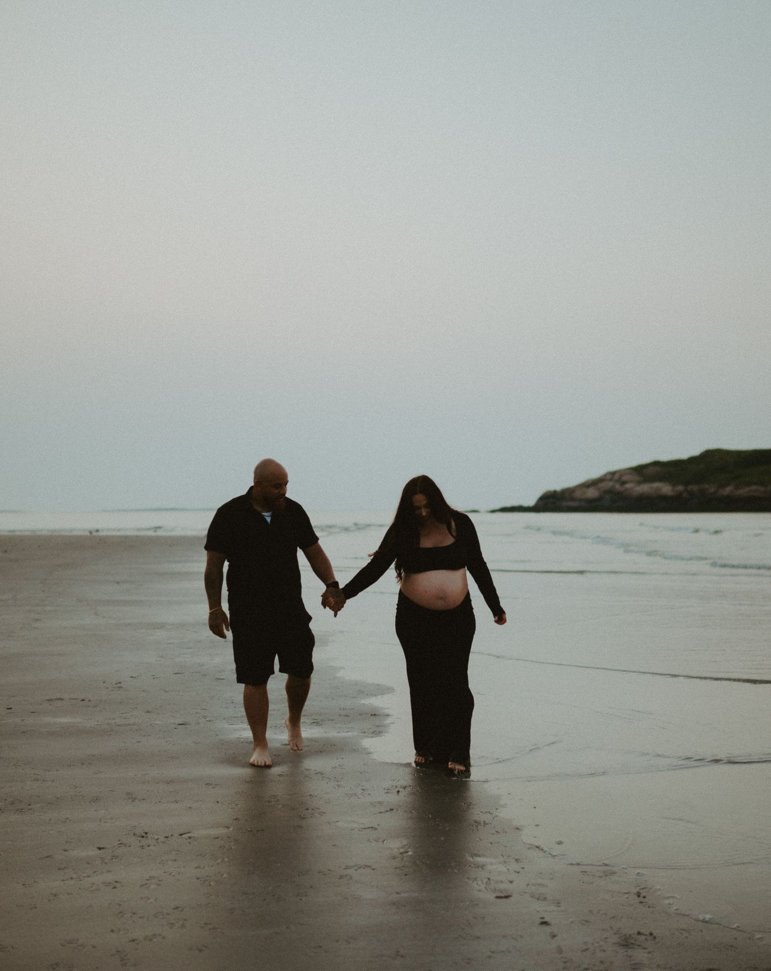 Couple holding hands walking barefoot on beach, pregnant woman in black outfit with partner.