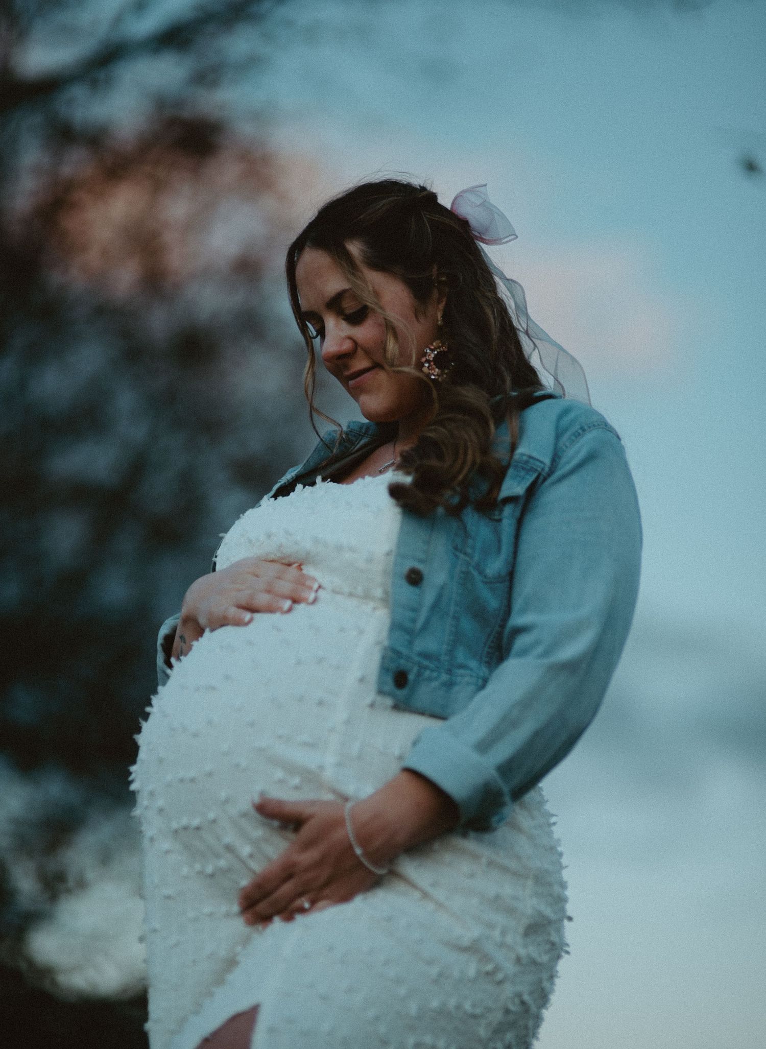 Pregnant woman in white textured dress and denim jacket gazes down at baby bump during outdoor maternity shoot.